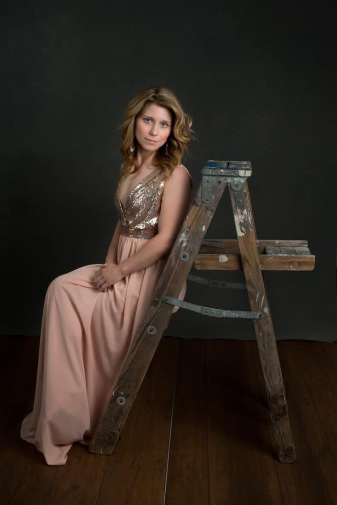 blonde model wearing a long pink sequin dress, sitting on a wood ladder in South Jersey portrait studio.