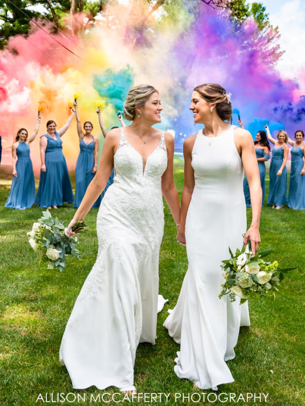 Two brides holding hands and walking in front of Rainbow Smoke Bombs at their Legacy Club at Woodcrest Wedding in