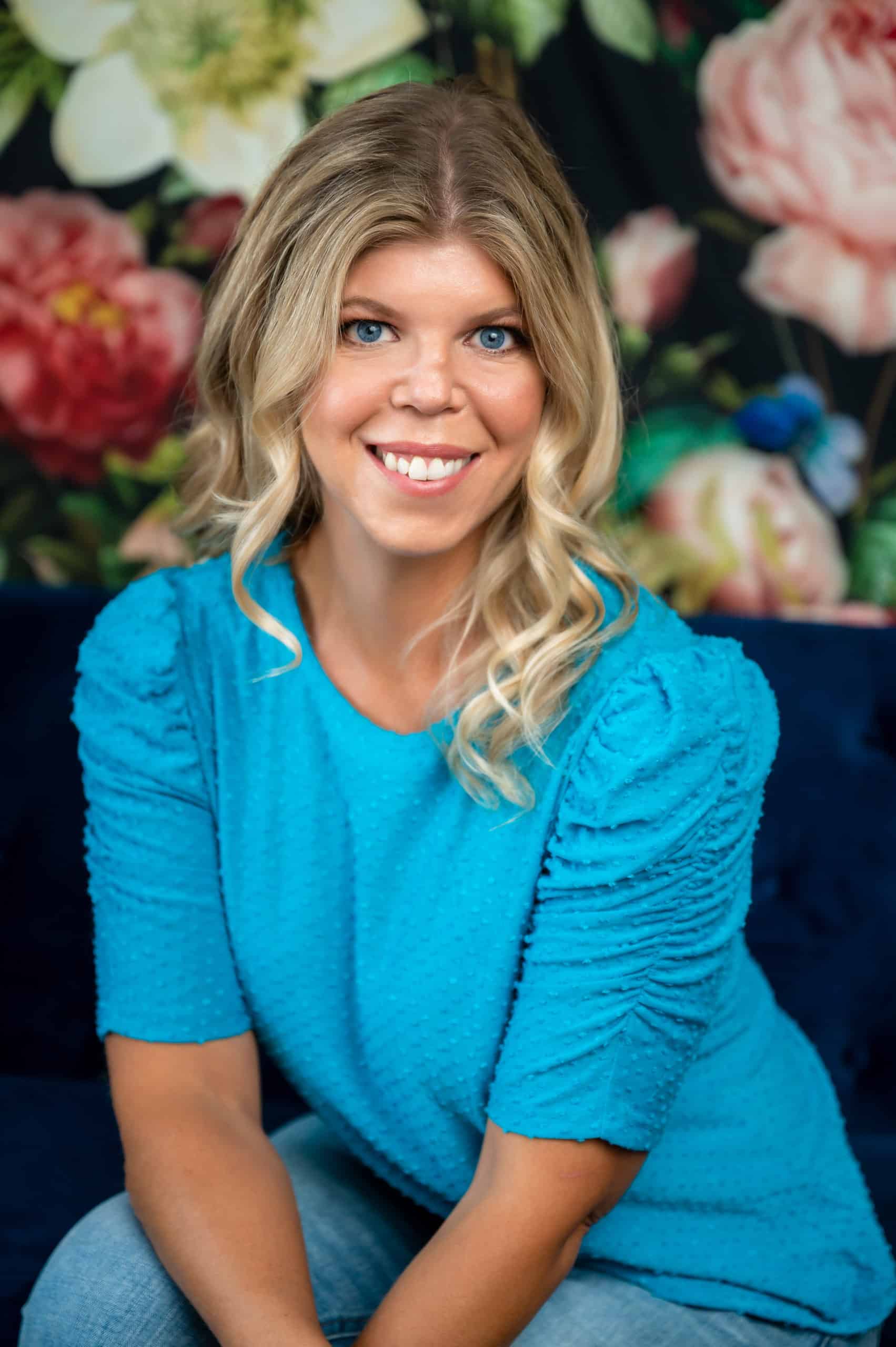 Headshot of a blonde woman with a blue shirt and blue eyes in front of a floral backdrop taken in a South Jersey headshot studio