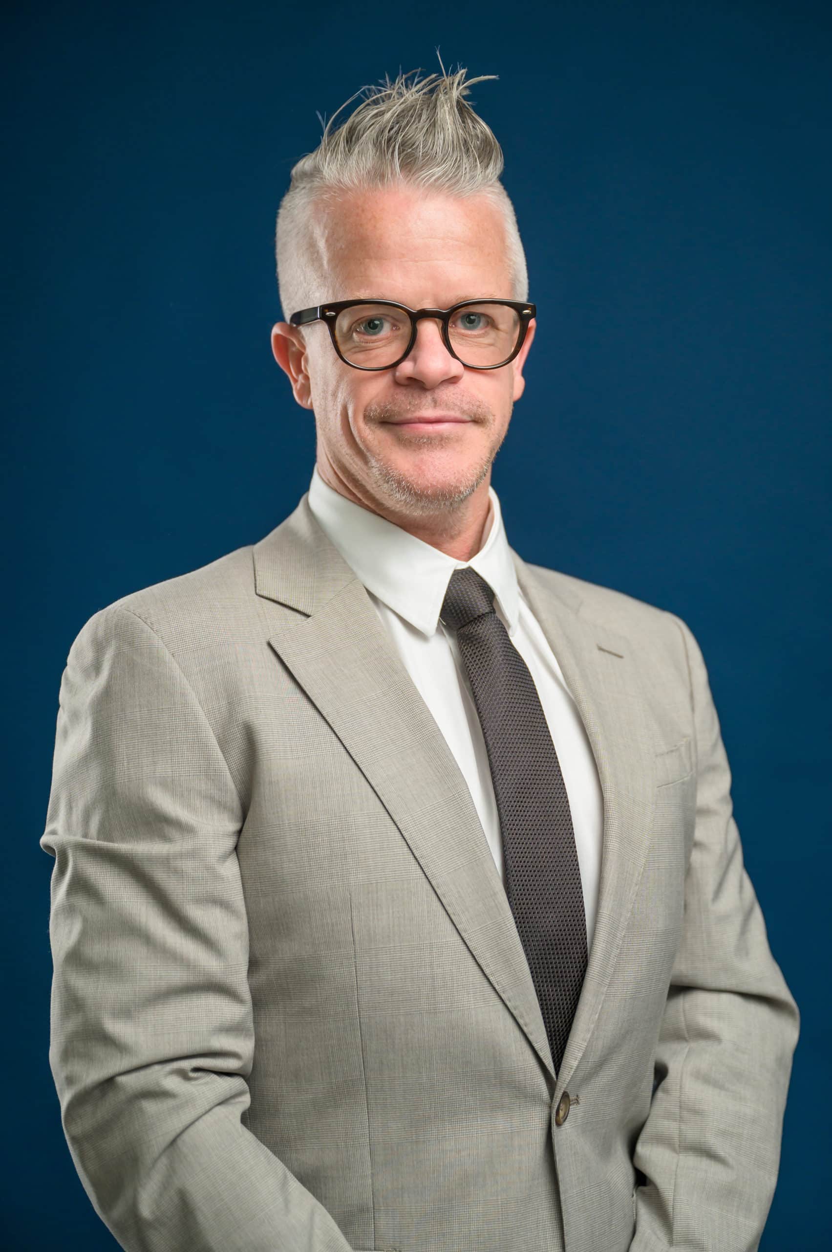 Professional headshot of a man wearing a tan suit wearing glasses in front of a blue backdrop