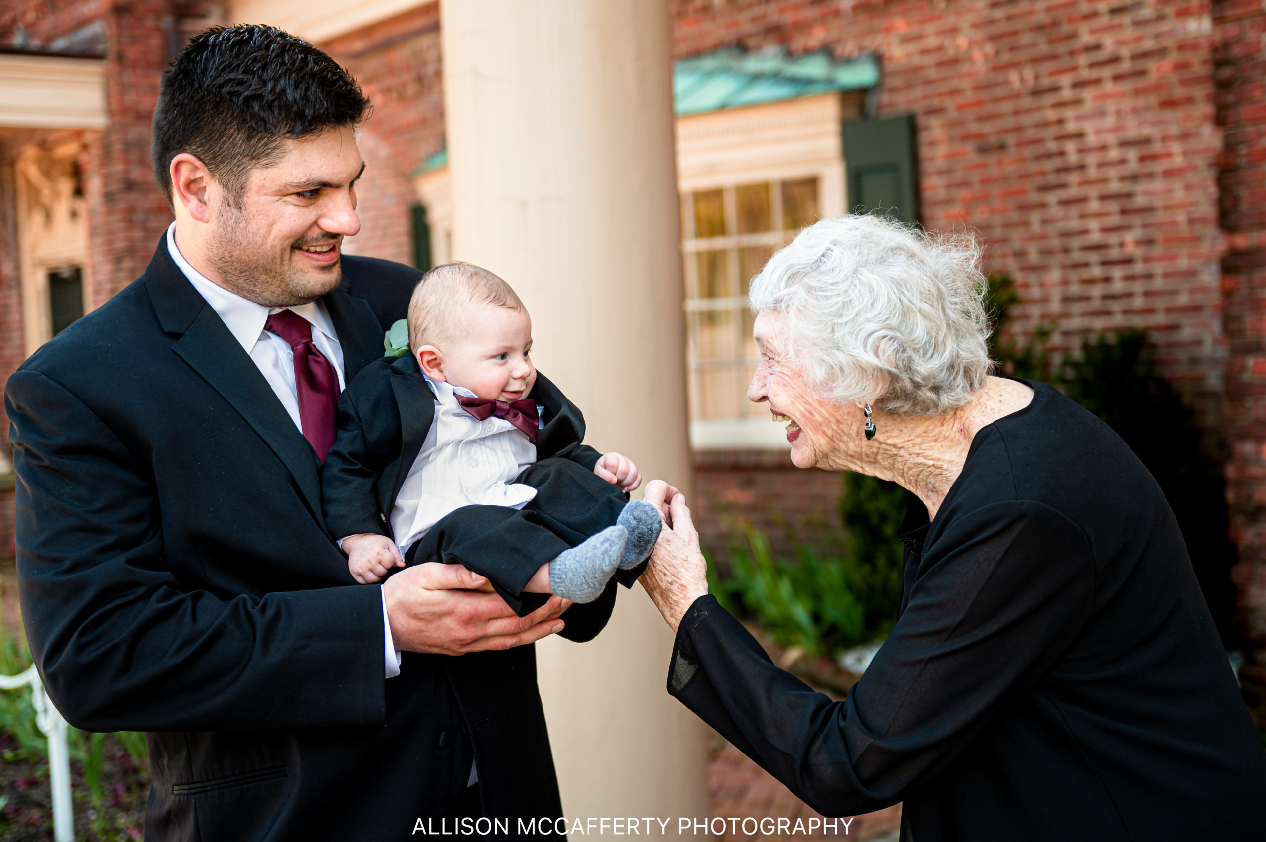 Wedding photo of a man holding his baby son wearing a suit and his grandmother is smiling at him in front of The Manor in West Orange NJ