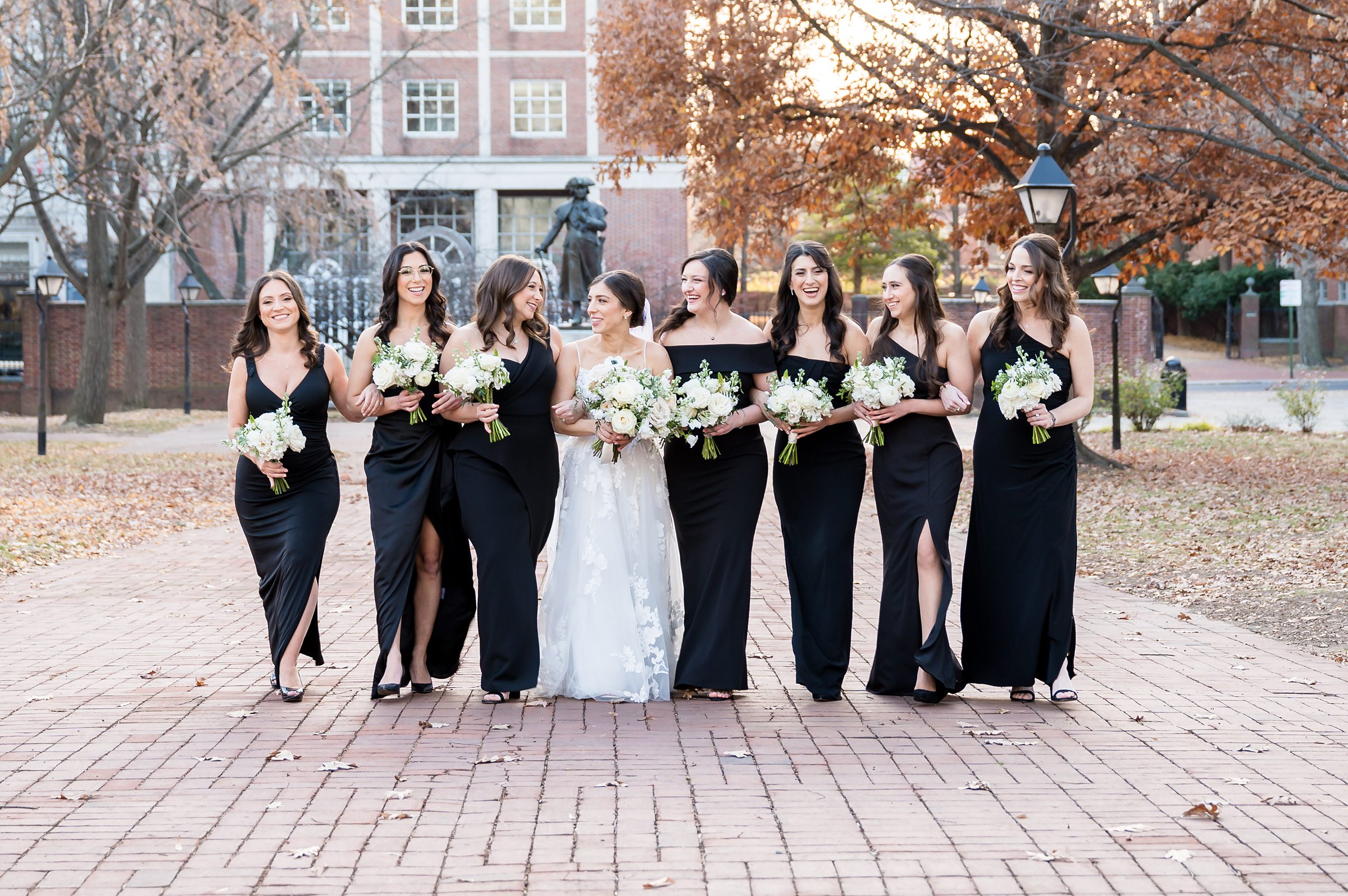 A bride and her bridesmaids in elegant black dresses pose on a brick walkway during a Lilah Events wedding.