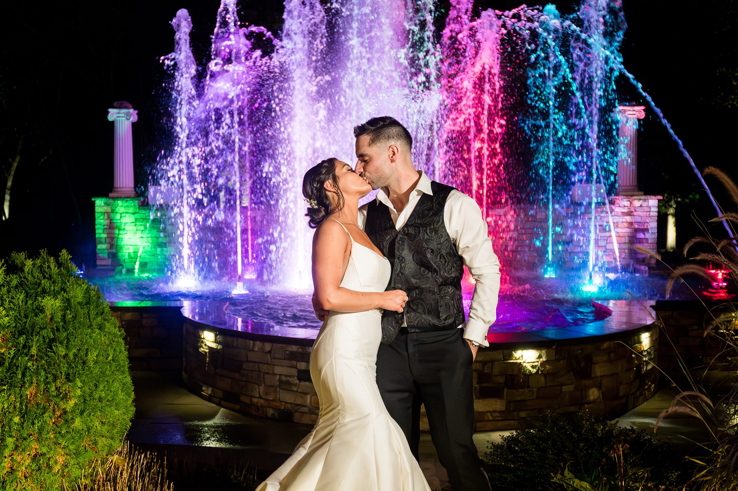 Bride and groom at their wedding standing in front of a fountain which is lit in several colors at Brigalias in Williamstown NJ