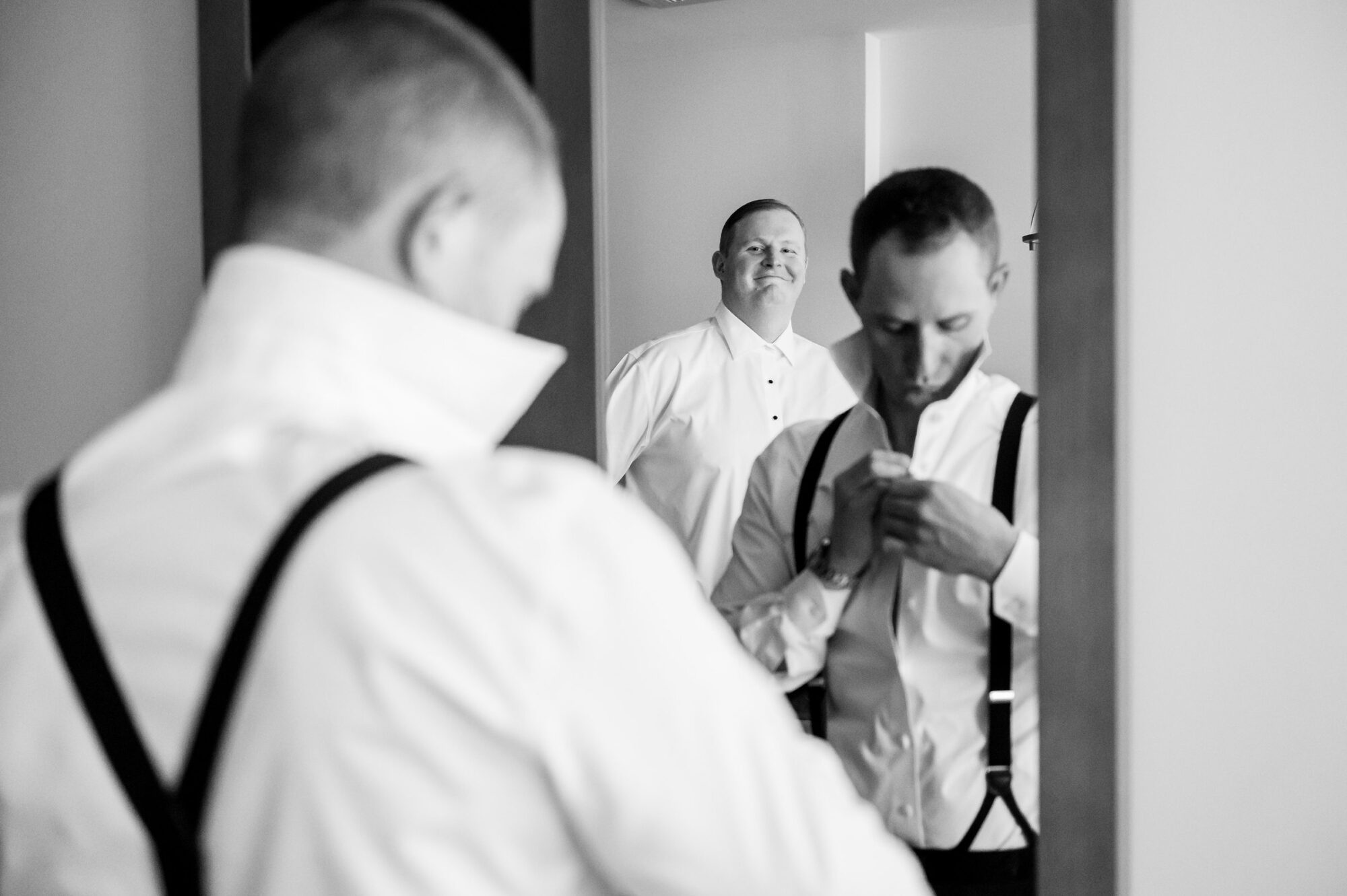 A man is putting on his suspenders in front of a mirror.