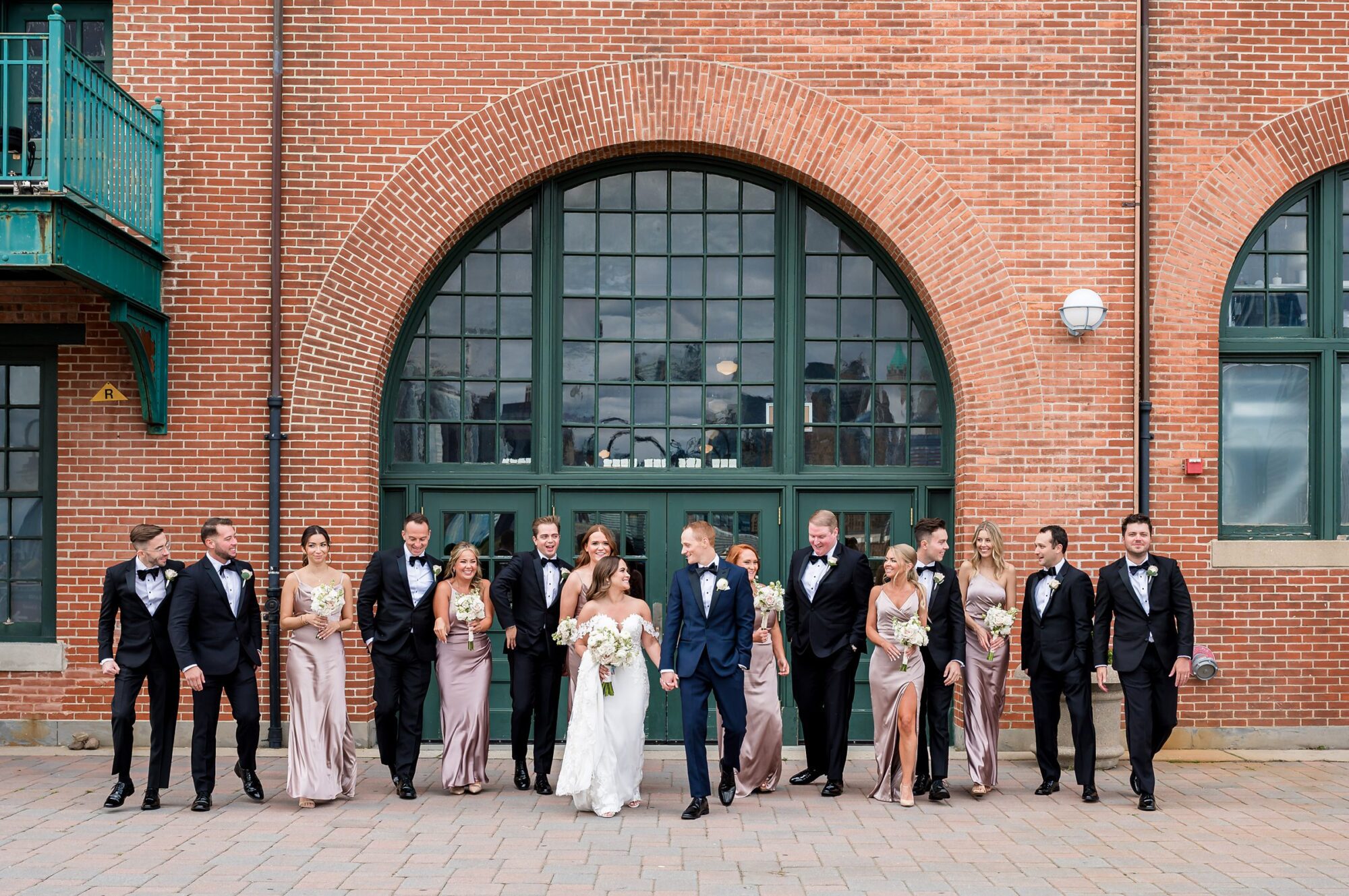 A group of bridesmaids and groomsmen pose in front of a brick building.