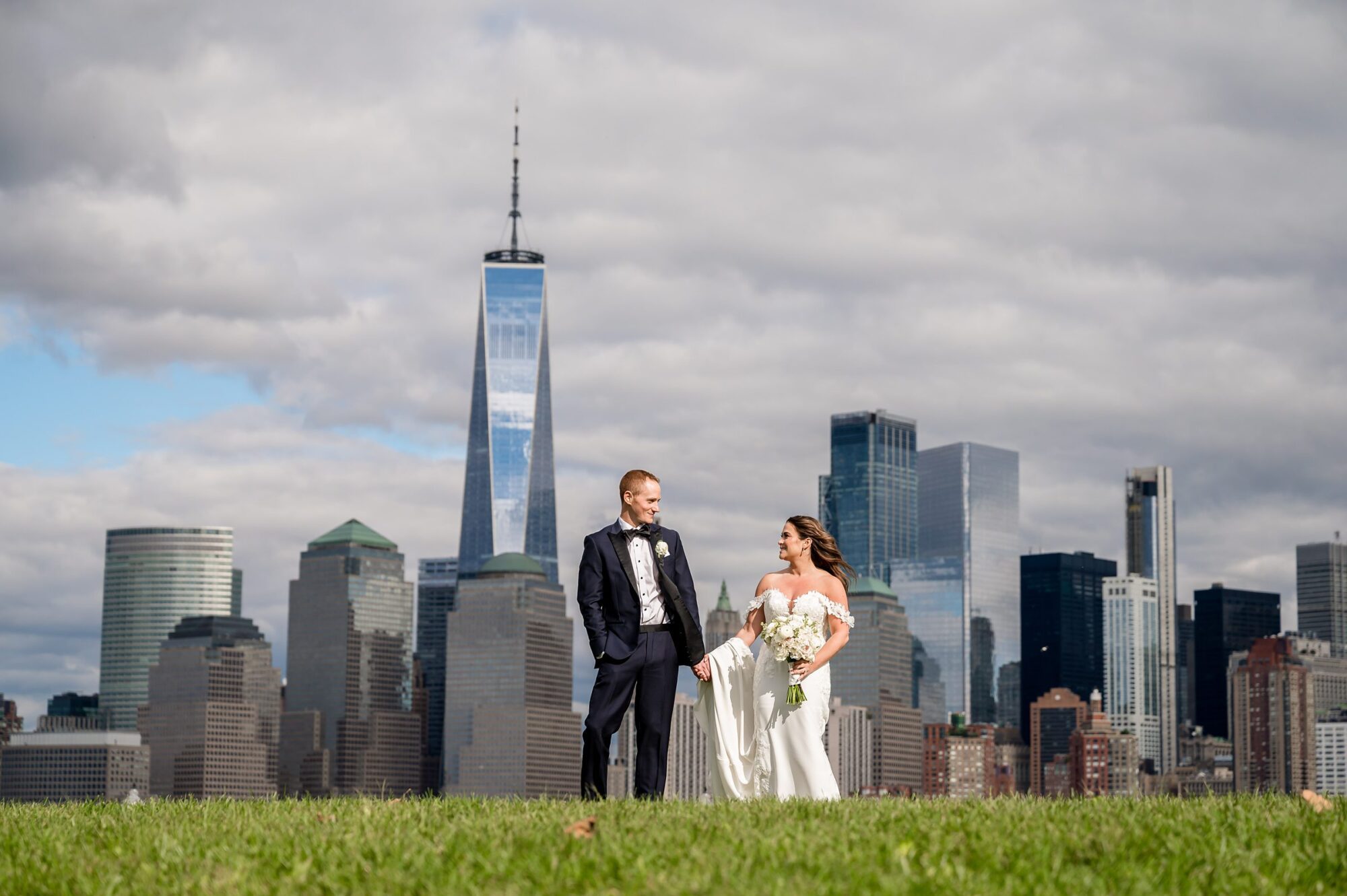 A bride and groom standing in the grass with the NYC skyline in the background.