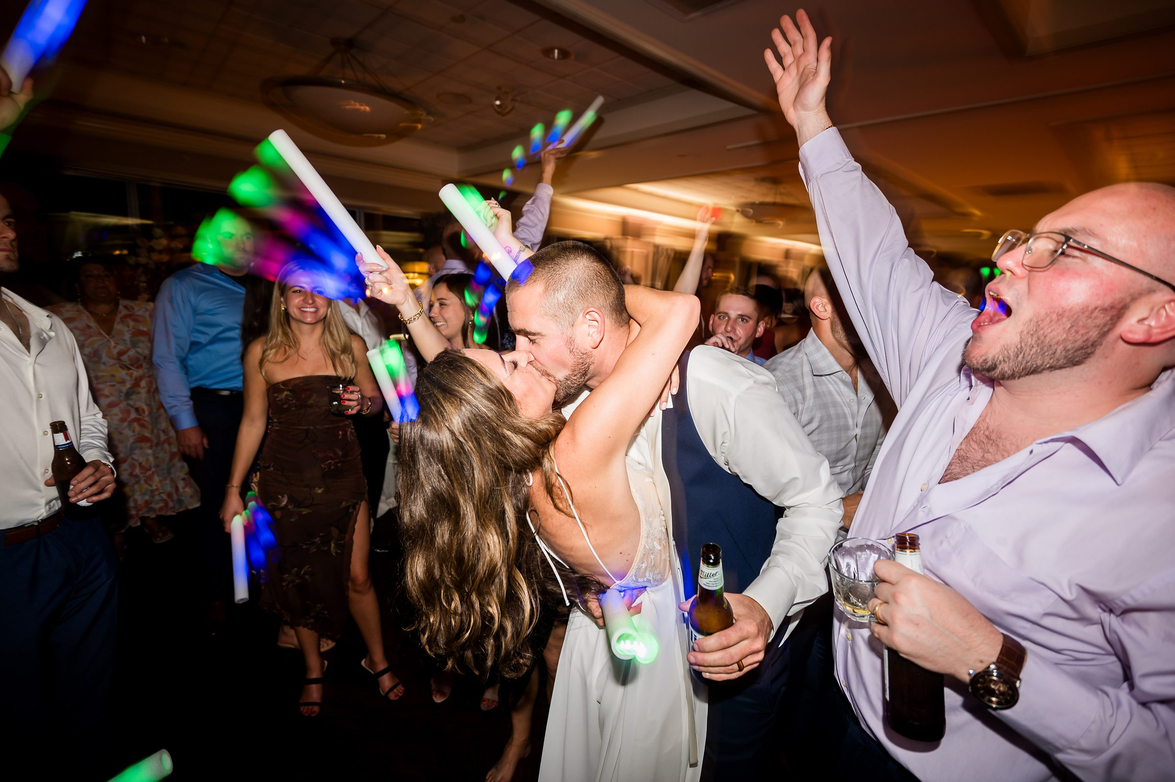 A couple is kissing on the dance floor during a lively party while surrounded by guests holding glowing sticks and a man to the right singing along with a drink in hand.