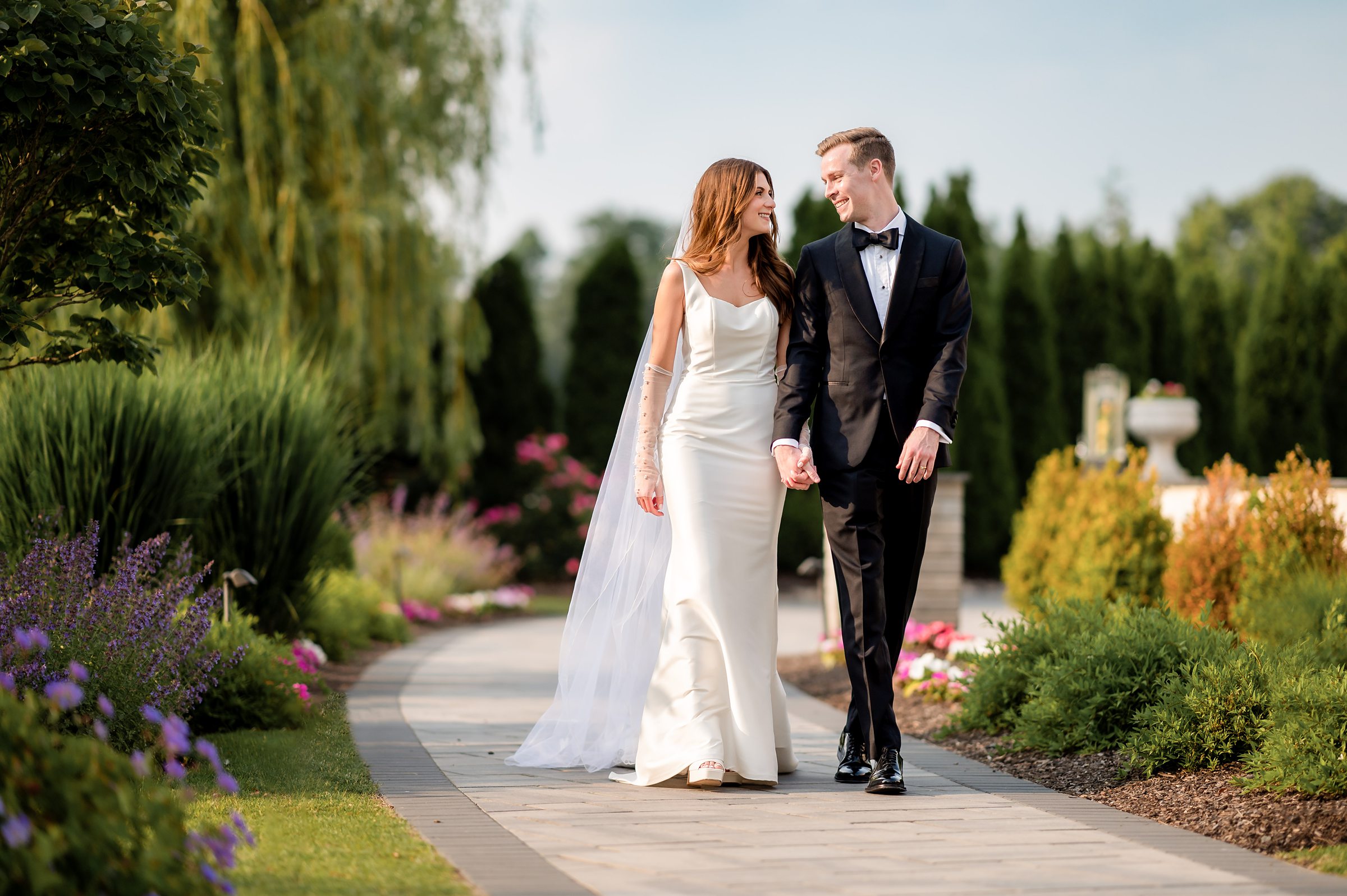 A couple in wedding attire walking hand in hand along a garden path on the grounds of The Mansion on Main Street in Voorhees NJ