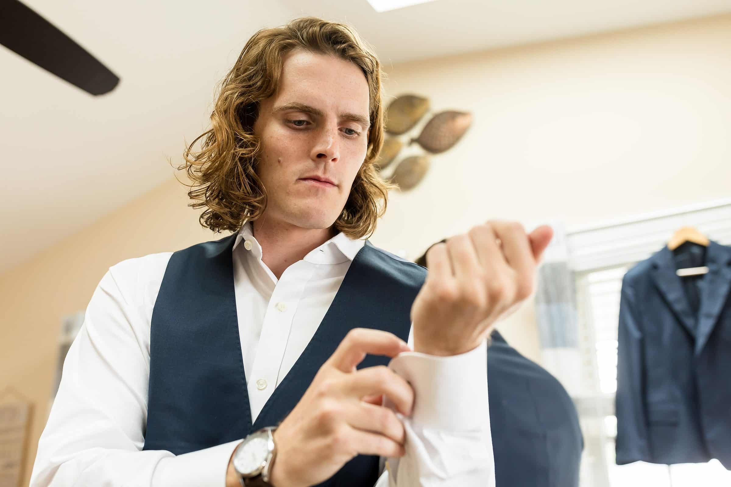 A man with long hair adjusts the cuff of his white dress shirt while wearing a navy vest. A suit jacket is hung in the background.