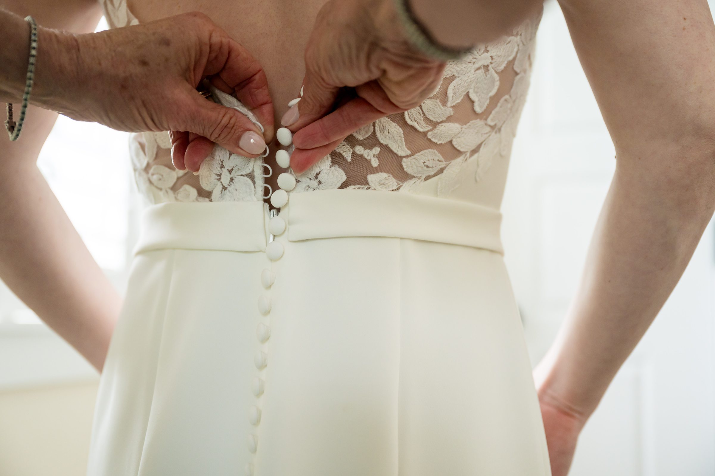 Close-up of a person buttoning the back of a white wedding dress adorned with lace.