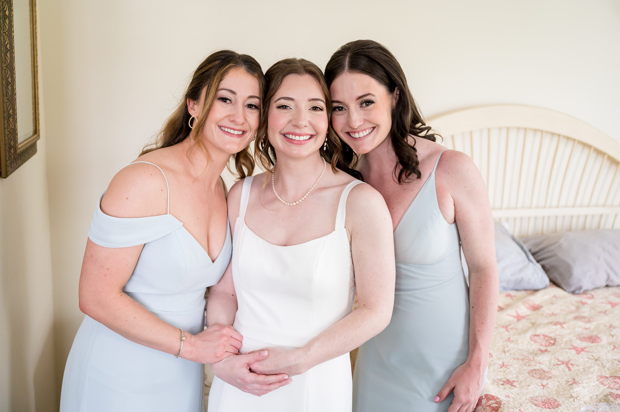 Three women pose together indoors. The woman in the center wears a white dress, and the women on either side wear light blue dresses. They are all smiling, with one bed visible in the background.