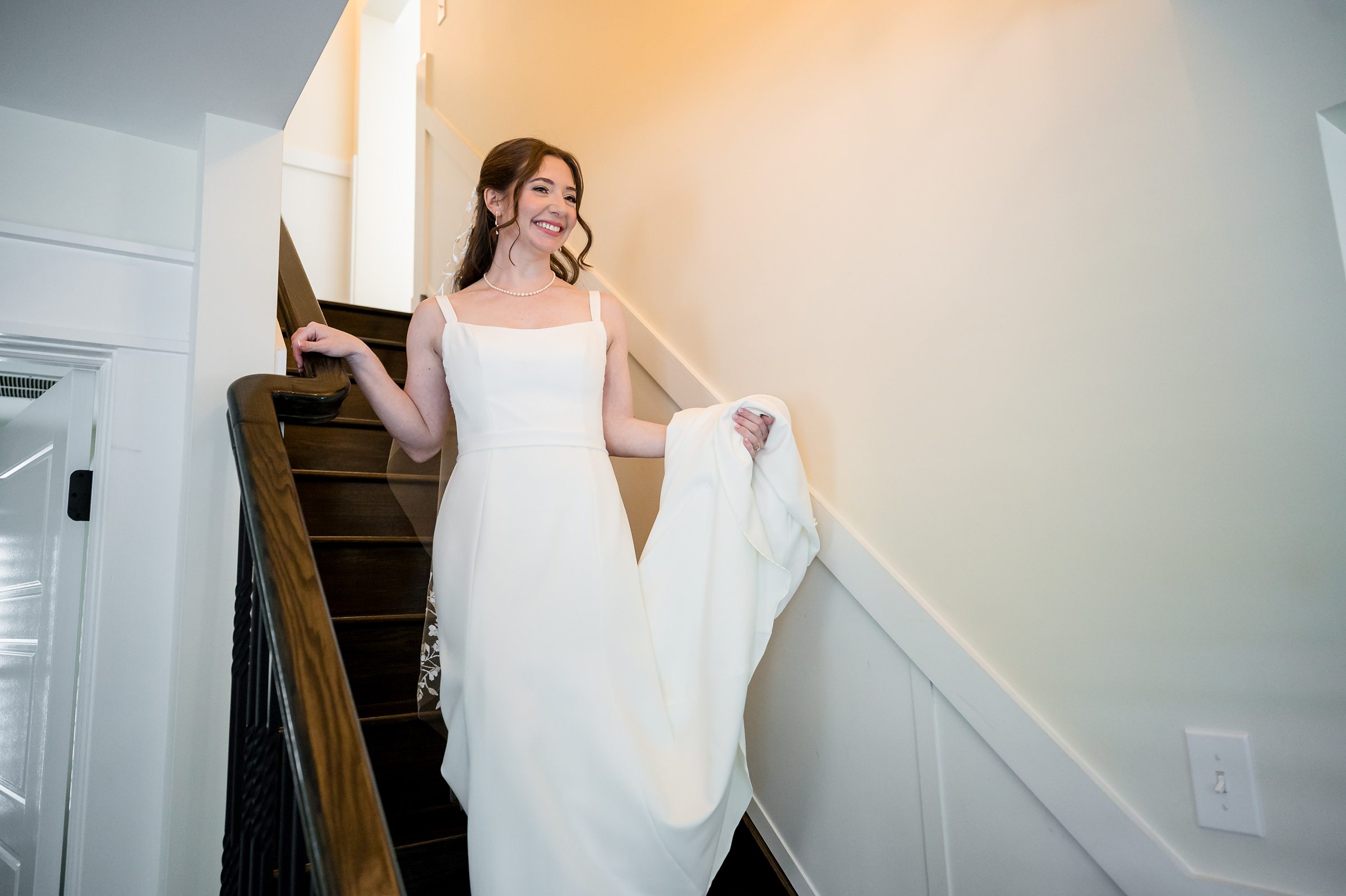 A woman in a white dress descends a wooden staircase with a slight smile.