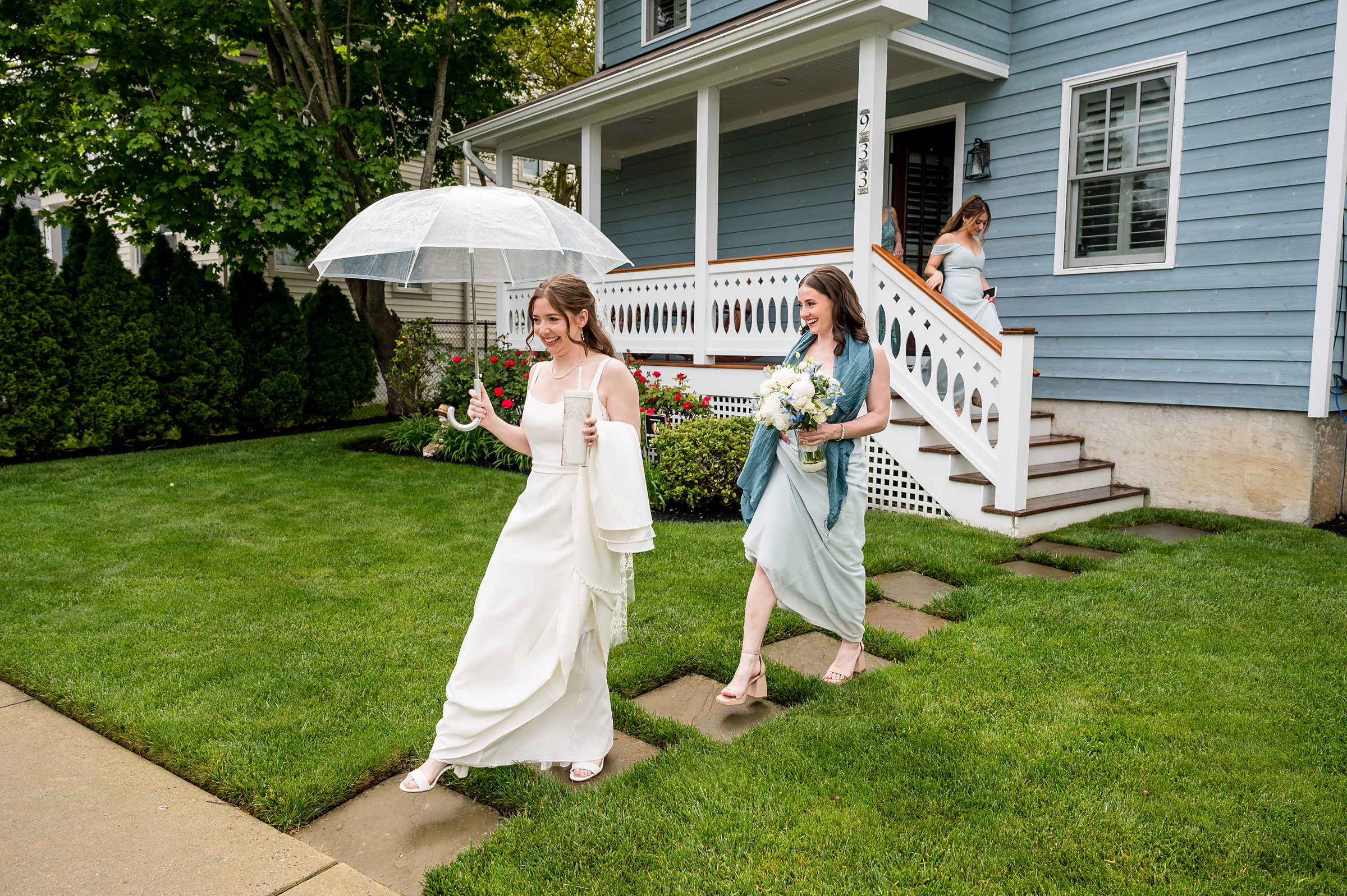 A bride holding an umbrella walks on a pathway beside a bridesmaid holding a bouquet, with another bridesmaid following down the stairs in front of a blue house.