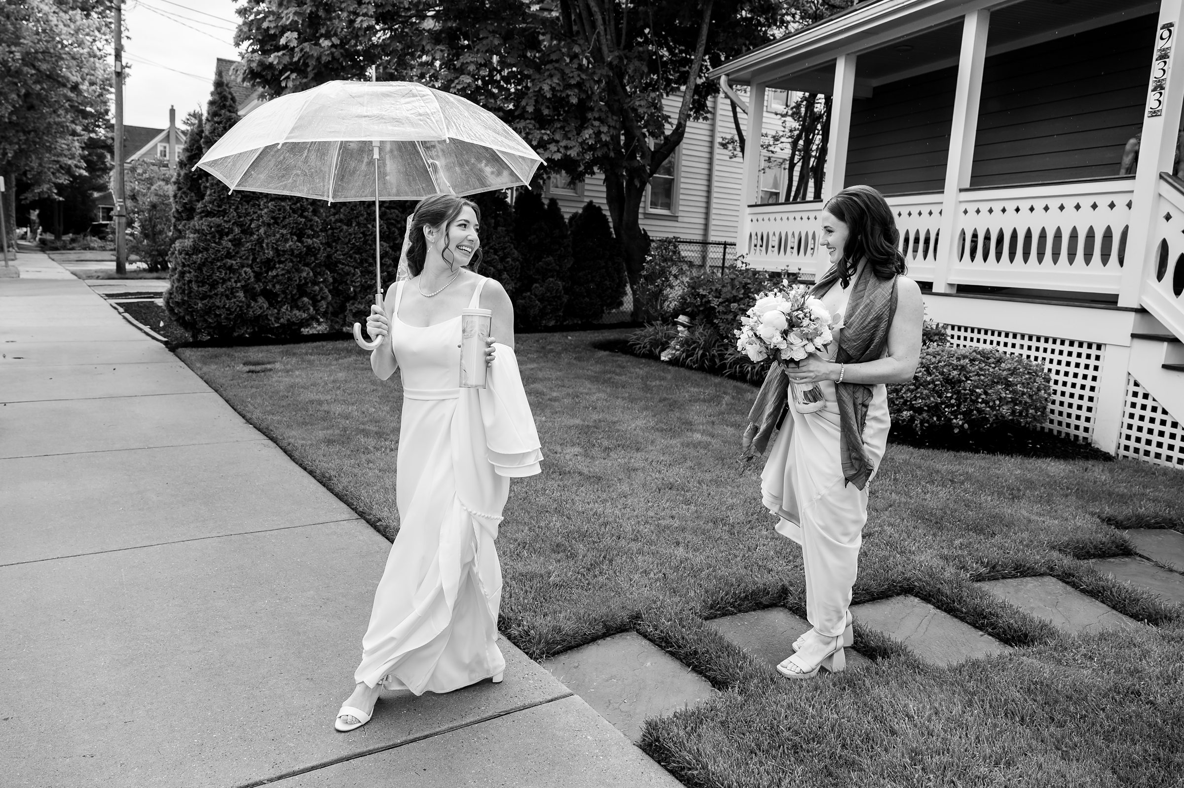 Two women wearing dresses walk near a house, one holding a transparent umbrella and a drink, and the other carrying a bouquet of flowers.