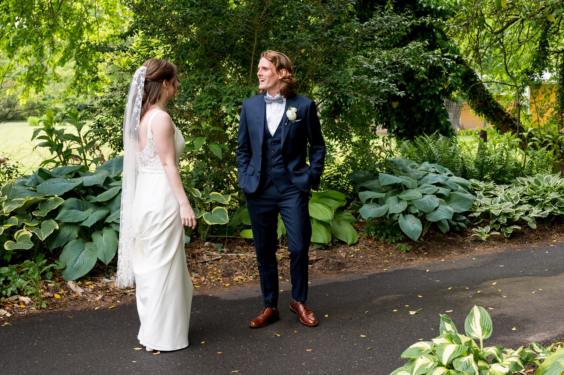 A bride in a white dress and veil and a groom in a navy suit stand facing each other outdoors on a path surrounded by greenery.