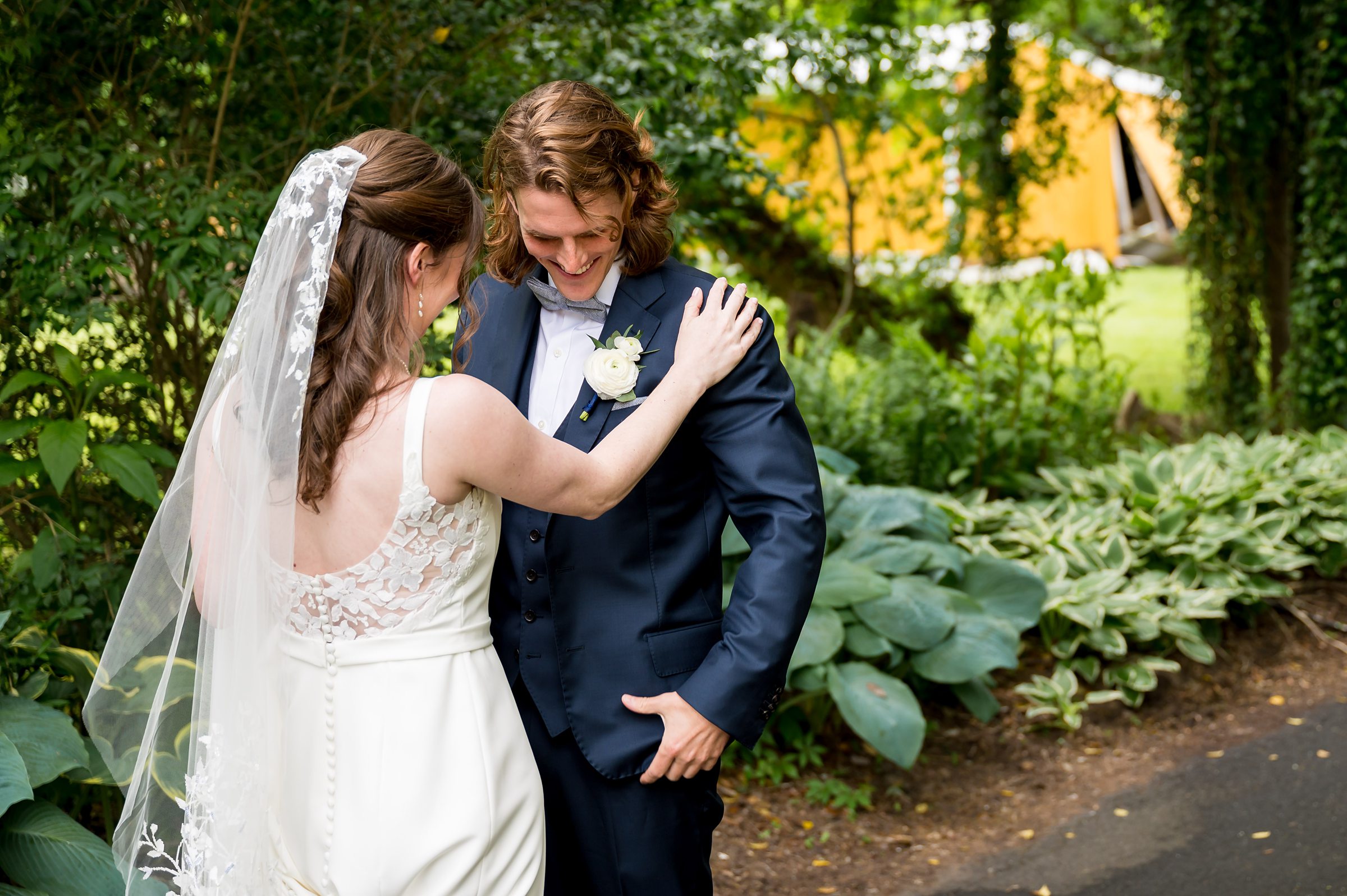 A bride and groom share a moment outdoors. The bride, in a white gown and veil, holds the groom's shoulders. The groom wears a dark suit and boutonniere, smiling and looking down. Lush greenery surrounds them.