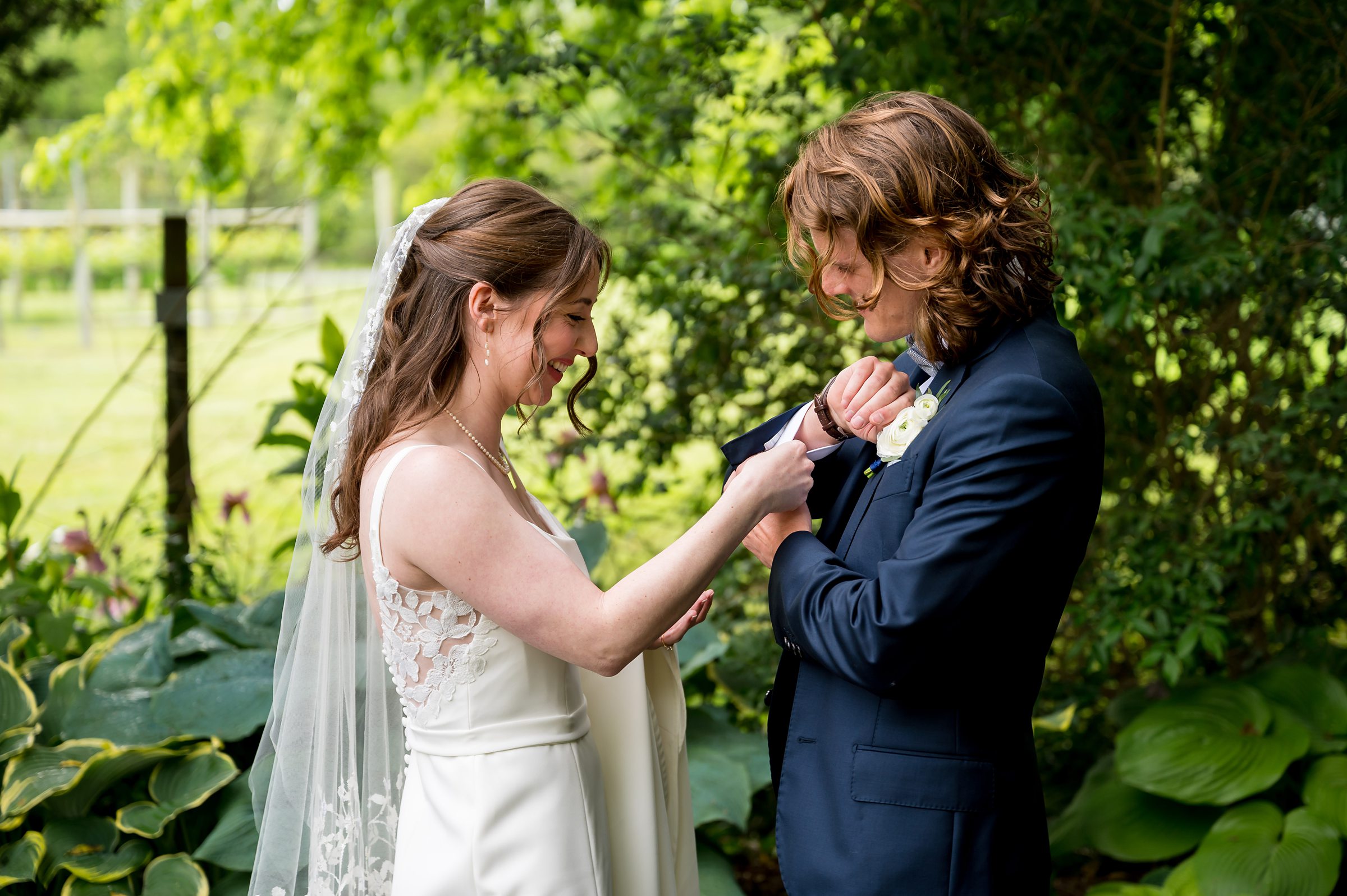 A bride adjusts the boutonniere on a groom's suit as they stand outdoors in a garden with lush greenery.