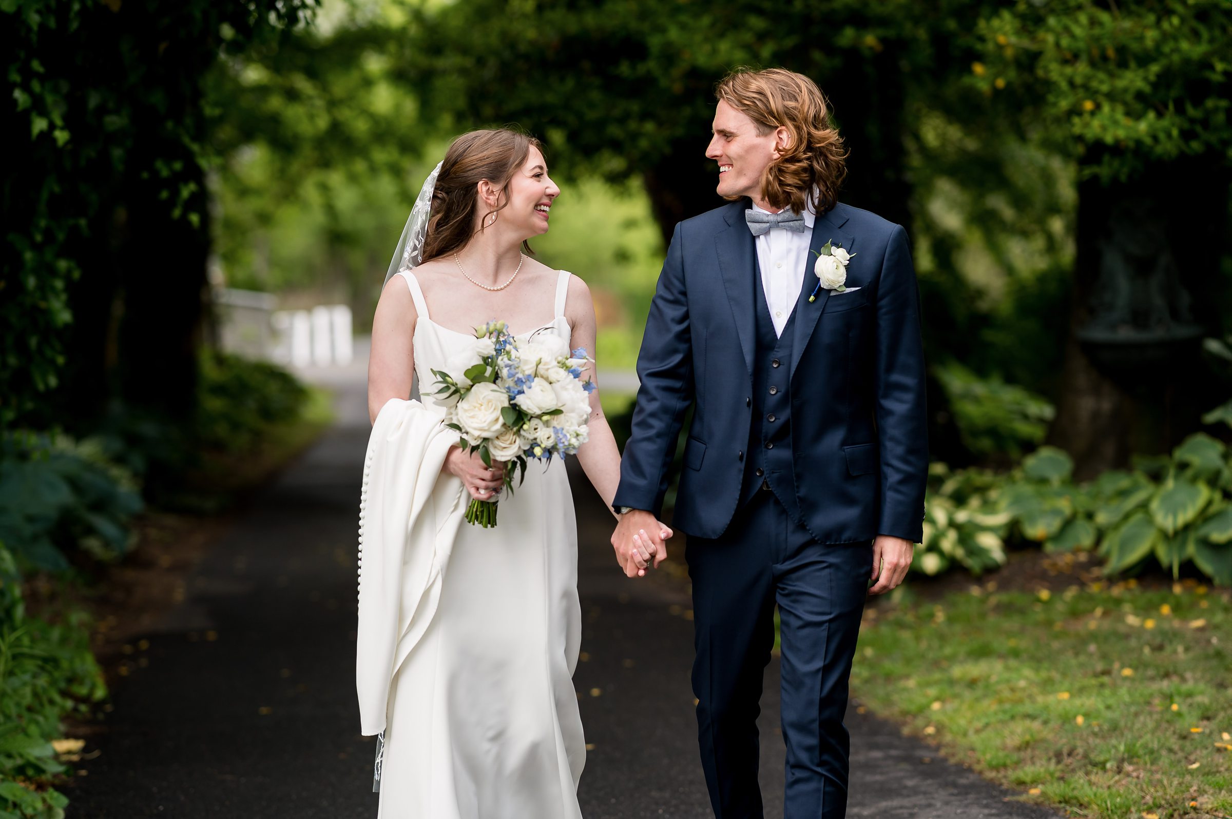 A bride and groom walk hand-in-hand on a pathway surrounded by greenery. The bride holds a bouquet and smiles at the groom, who wears a dark suit and bow tie. Both appear happy and relaxed.