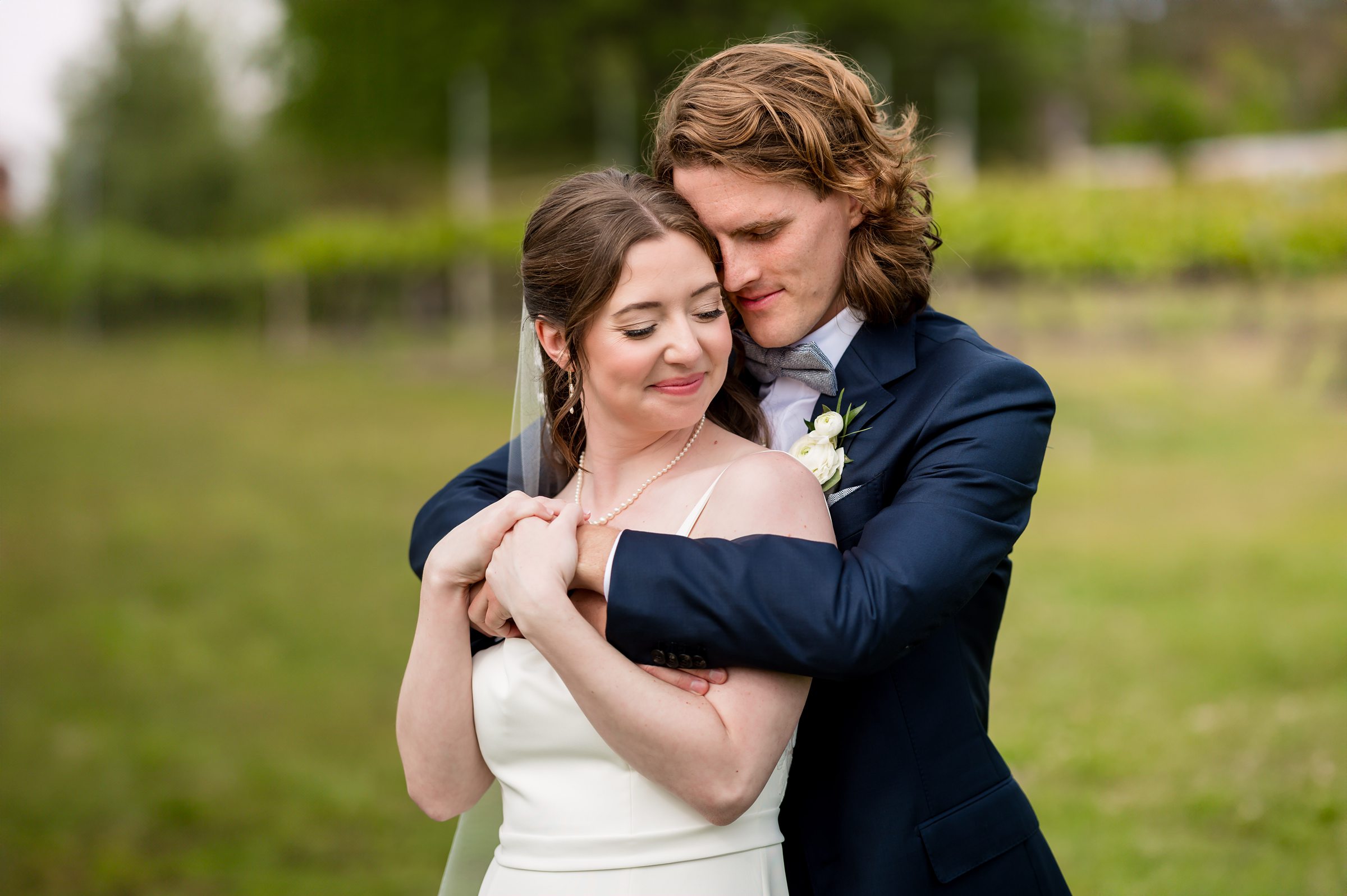 A bride and groom stand outdoors, with the groom hugging the bride from behind. The bride wears a white dress and the groom wears a dark suit. They both have their eyes closed, smiling softly.