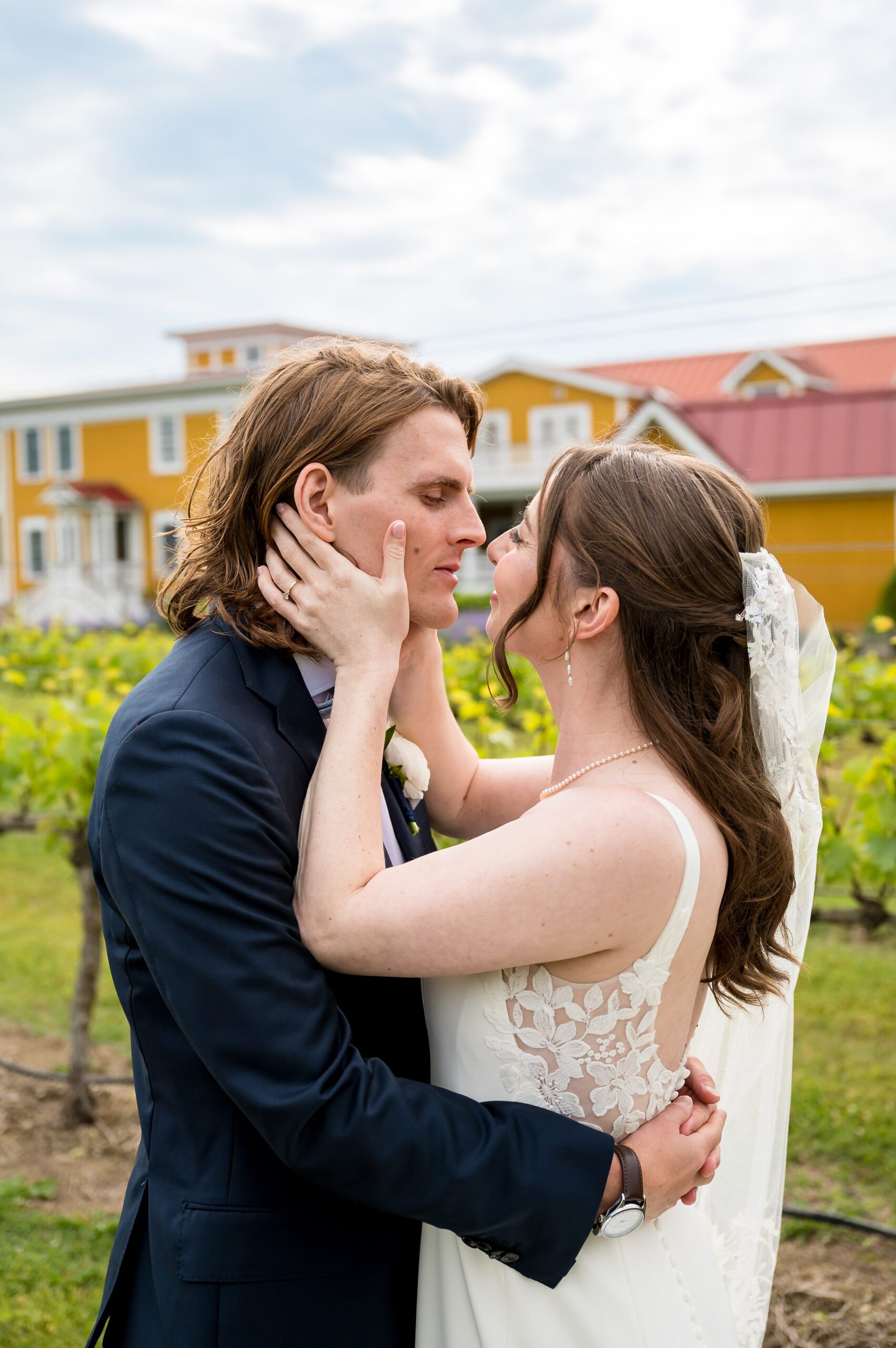 A bride and groom share an intimate moment, standing close and touching foreheads, against the backdrop of a building and greenery.