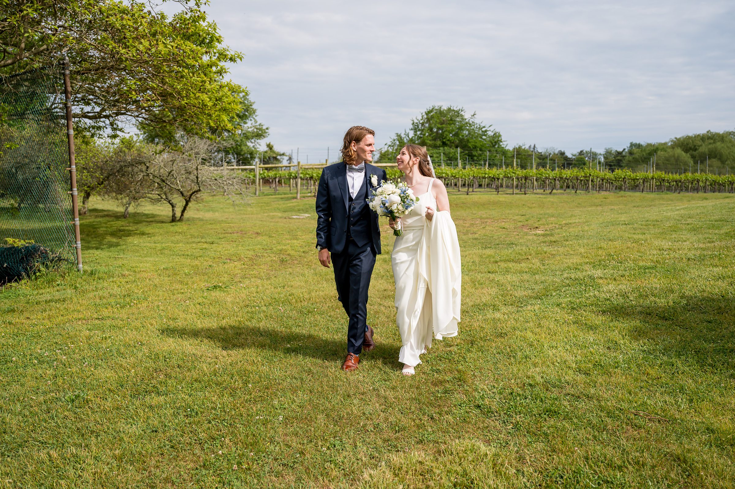 A bride and groom walk together on a grassy field. The bride carries a bouquet of flowers and wears a white wedding dress, while the groom wears a dark suit. Trees and greenery are visible in the background.