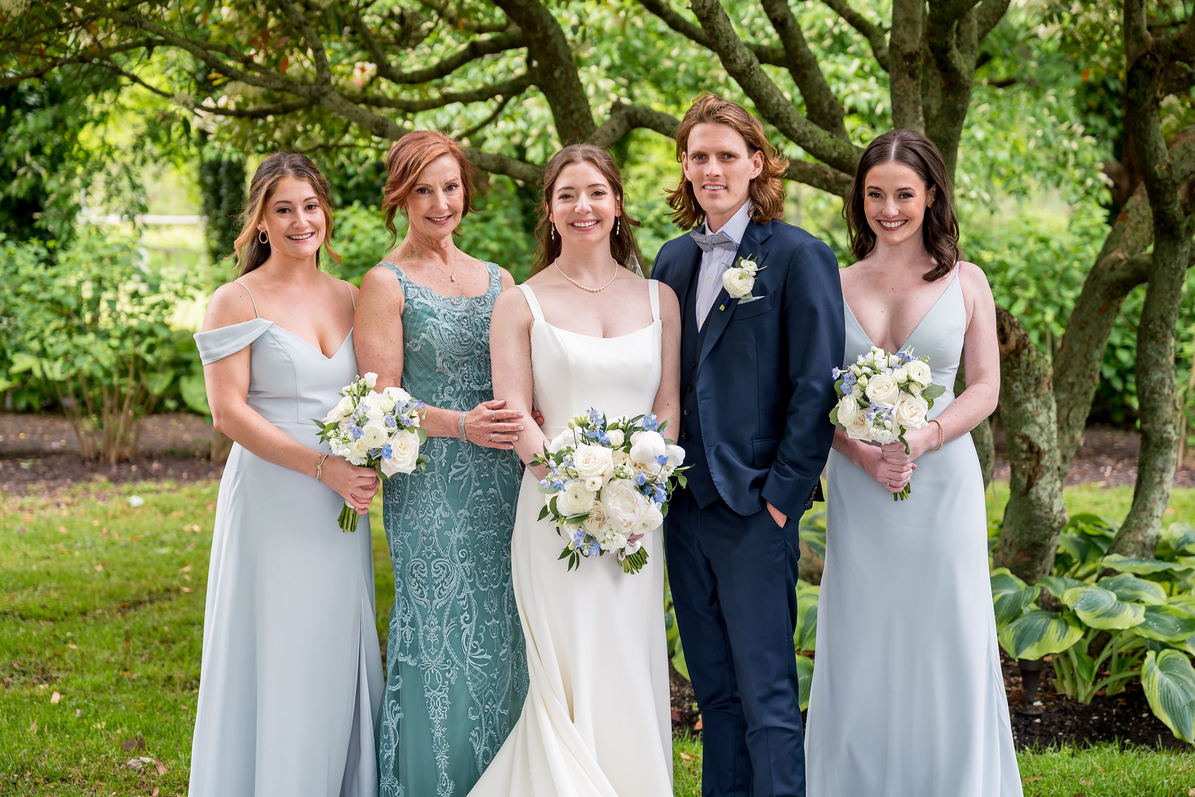 A group of five people, dressed in formal attire, are standing outdoors. Three women in light blue dresses and one man in a suit flank a woman in a white wedding dress holding a bouquet.