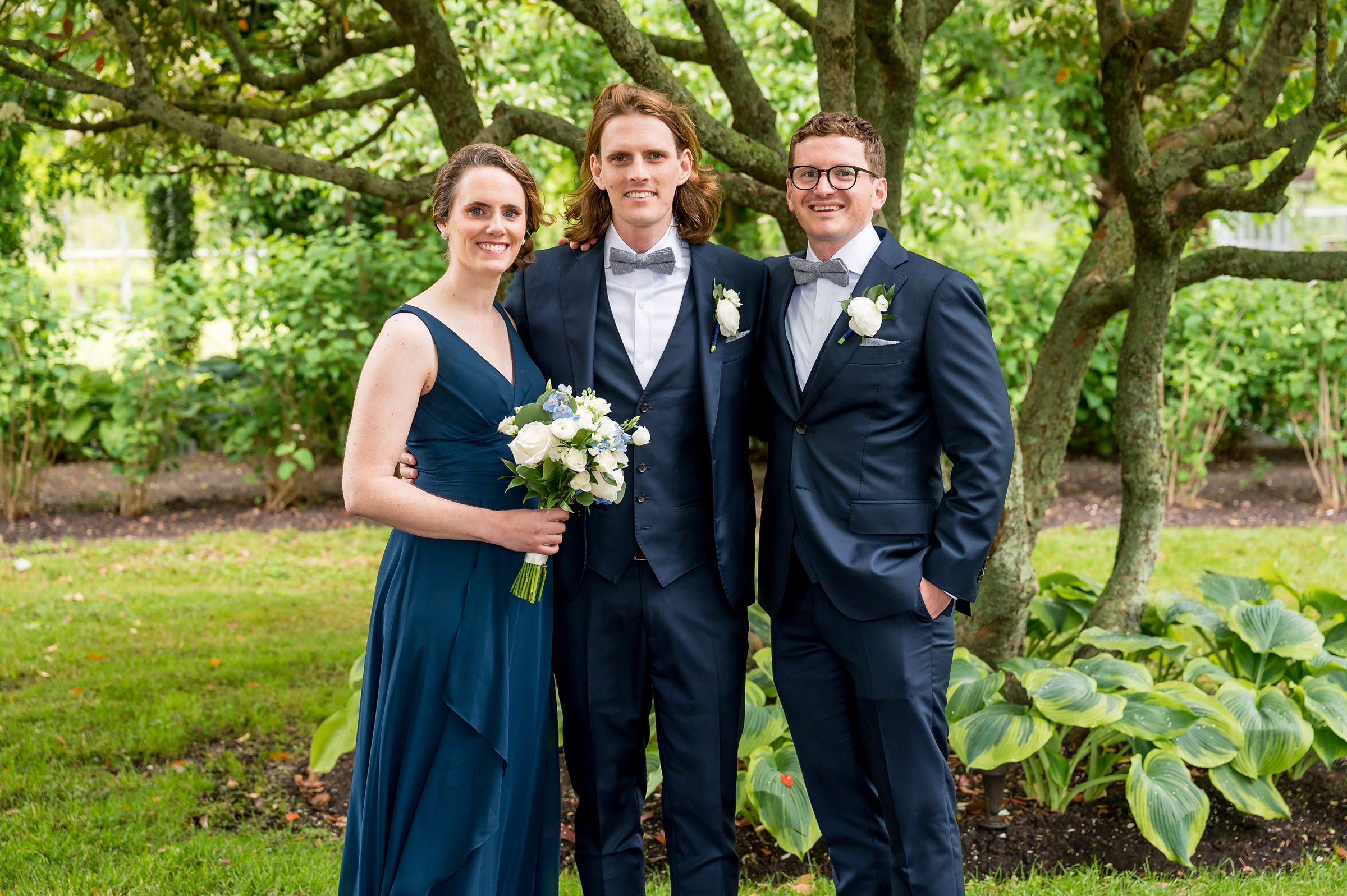 Three people dressed formally stand together outdoors, with two men in tuxedos and a woman in a navy dress holding a bouquet. Trees and greenery form the background.