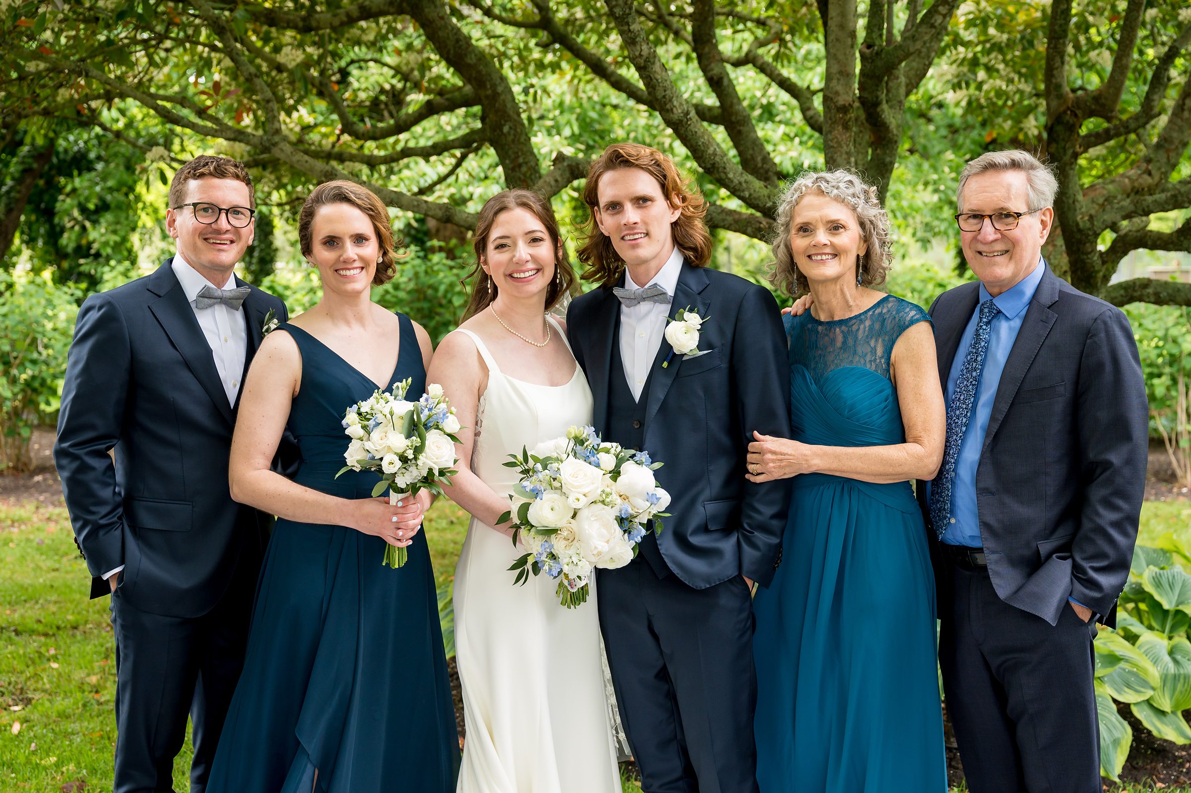 A wedding party poses outdoors in formal attire, including three men in suits and three women in teal dresses, with two holding bouquets. They stand in front of a tree with green foliage.