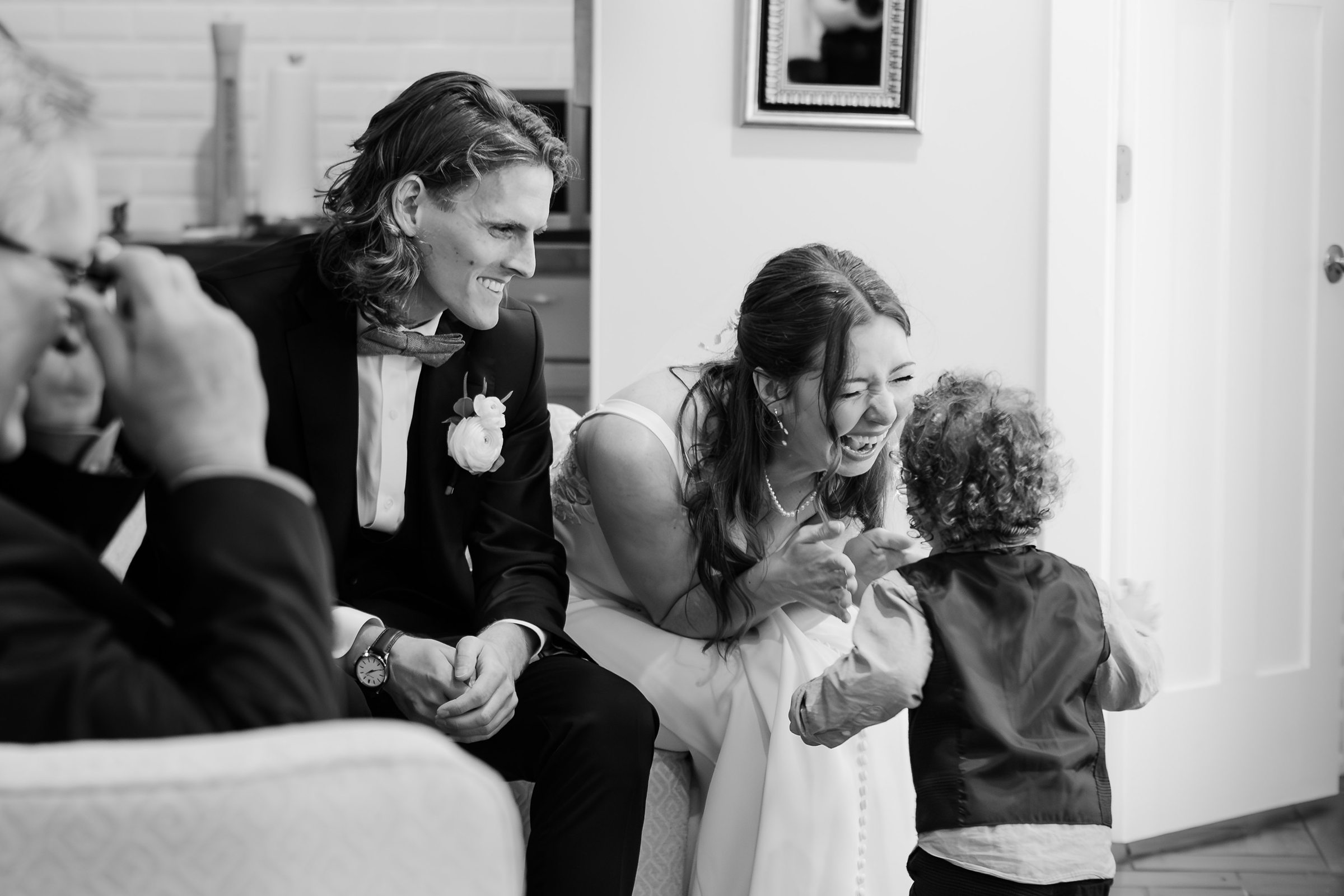 A bride laughs joyfully while interacting with a small child. The groom, seated beside her, smiles and watches. They are in an indoor setting with a mirror and door in the background.
