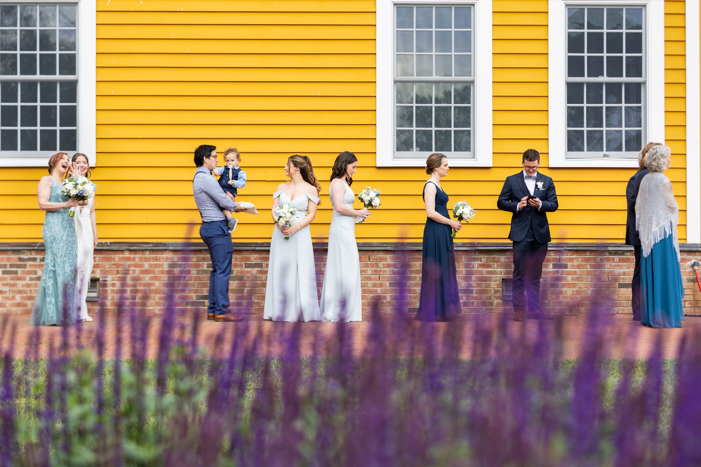 A group of people dressed formally, including bridesmaids and groomsmen, stand in front of a yellow building with white-framed windows, holding bouquets and engaging in various activities.