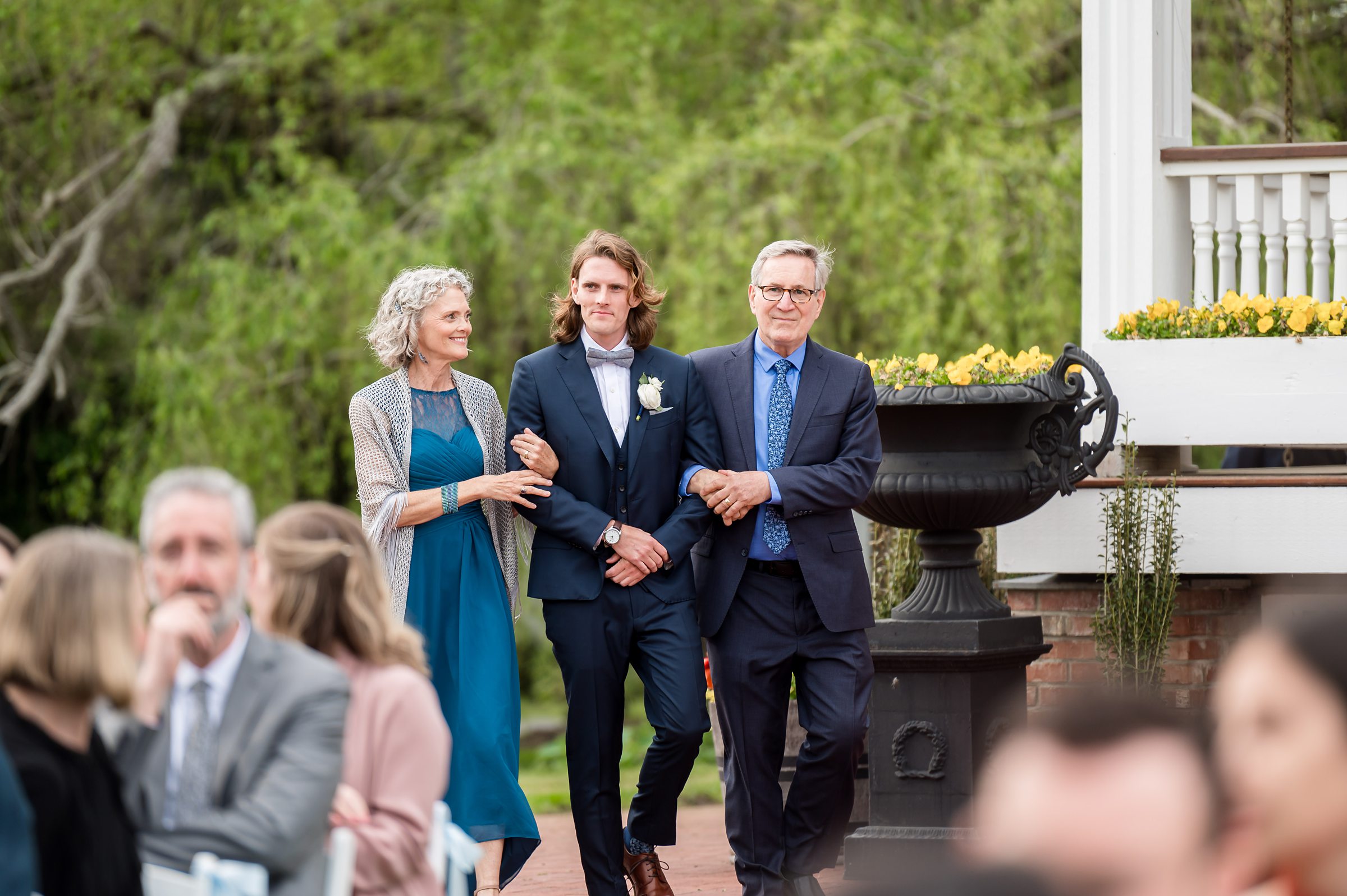 A groom walks down an outdoor aisle arm-in-arm with a woman and a man, surrounded by seated guests in a garden setting.