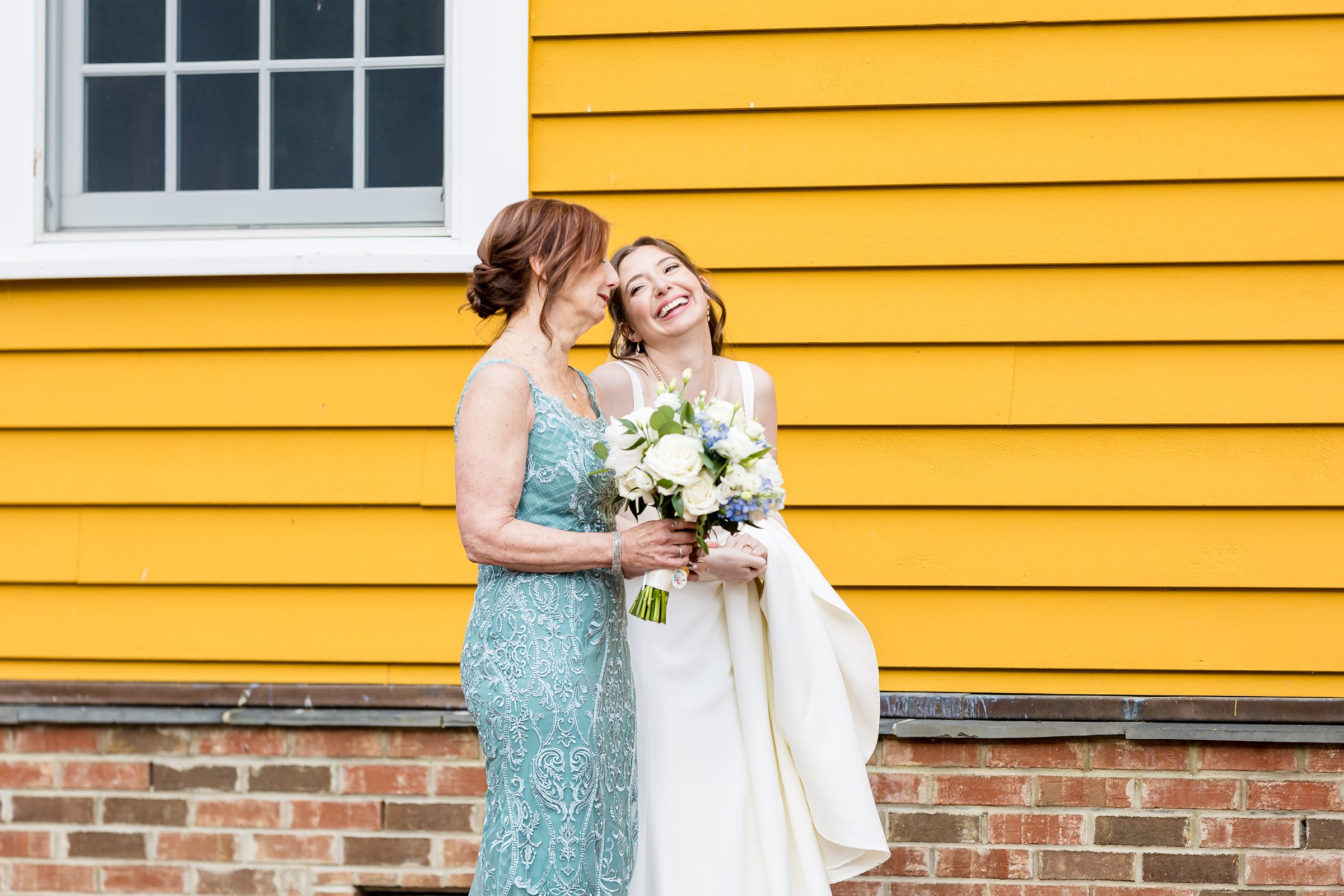 A woman in a green dress holds a bouquet while standing next to a smiling bride in a white dress in front of a yellow building.