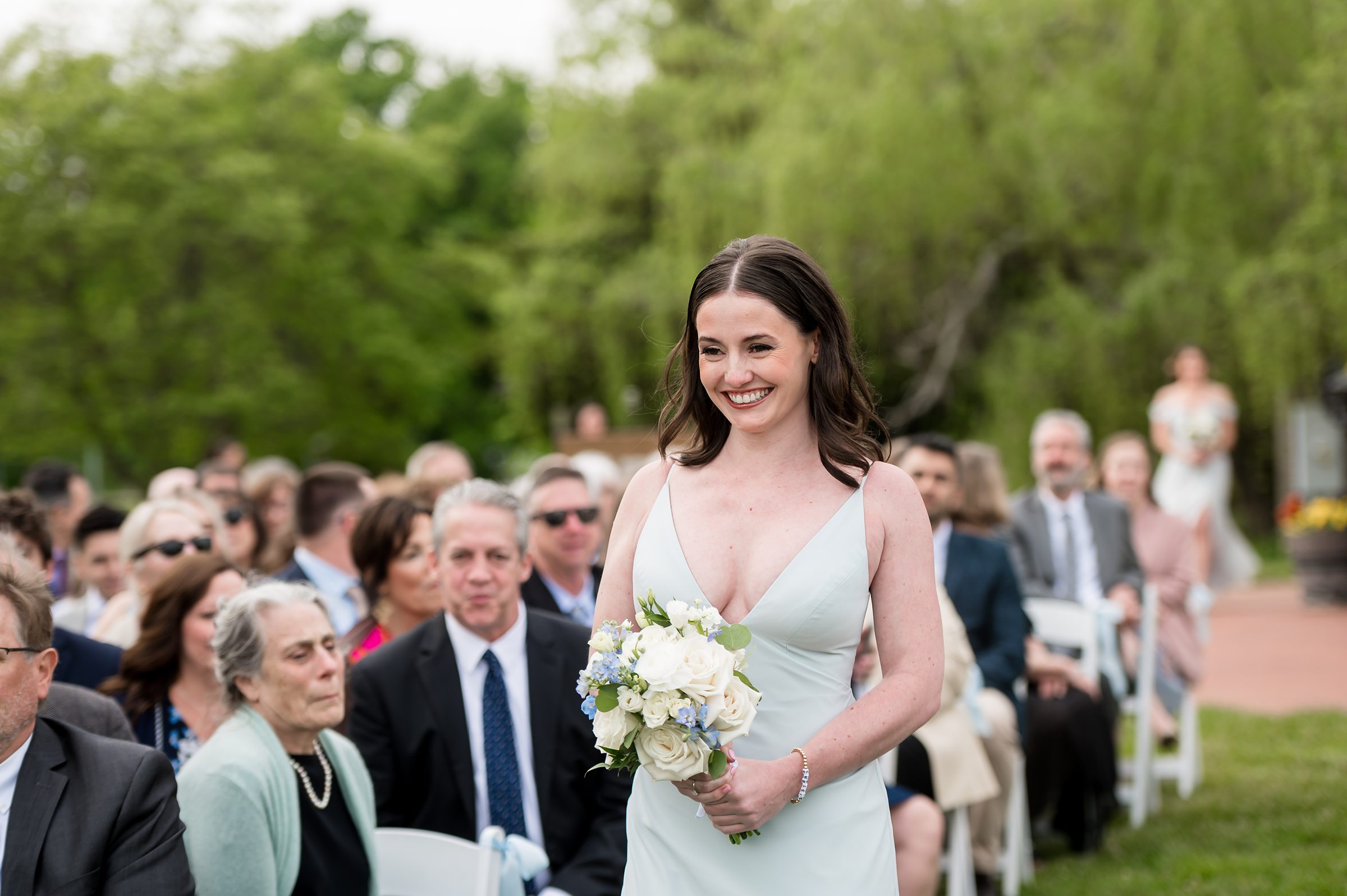 A woman in a light blue dress, holding a bouquet of flowers, walks down an outdoor aisle with seated guests on either side, during a daytime event. Trees are visible in the background.