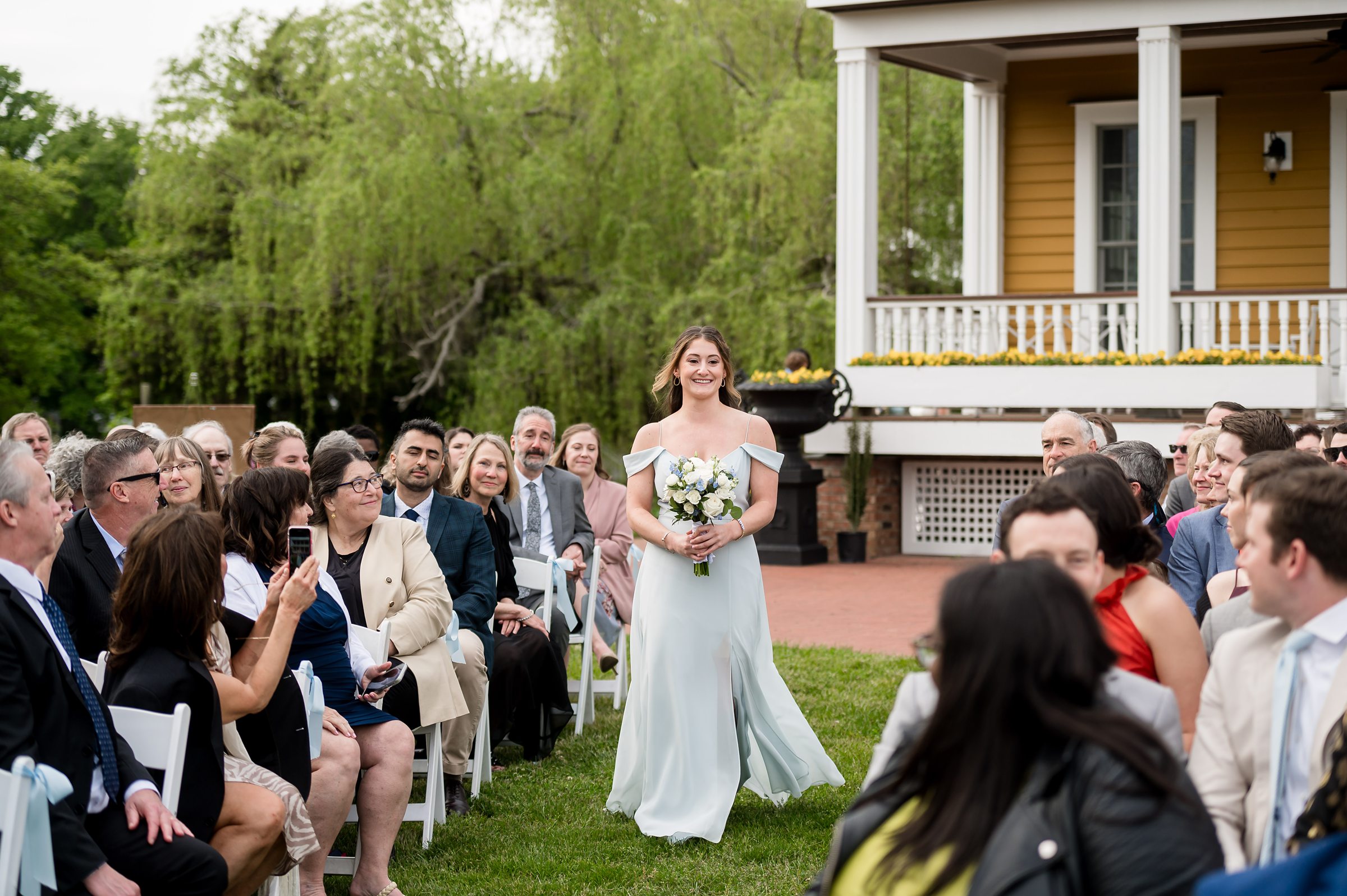 A woman in a light blue dress walks down an outdoor aisle holding a bouquet of flowers, with seated guests on either side, during what appears to be a wedding ceremony.