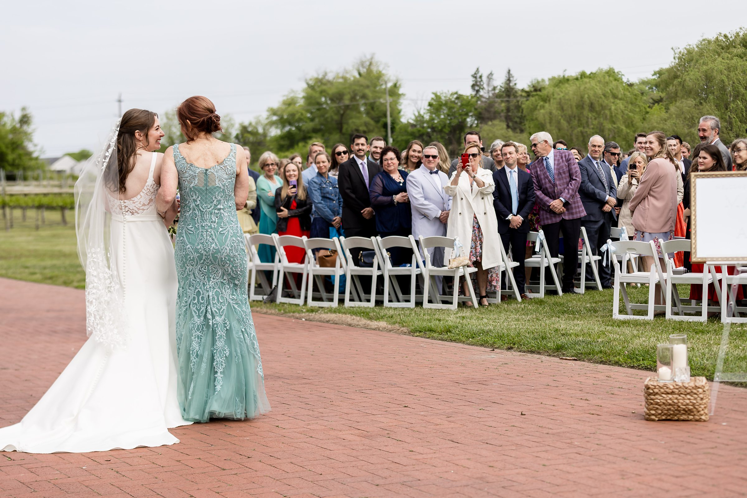 A bride in a white dress walks with another woman in a teal dress down an outdoor aisle as guests seated on white chairs look on.