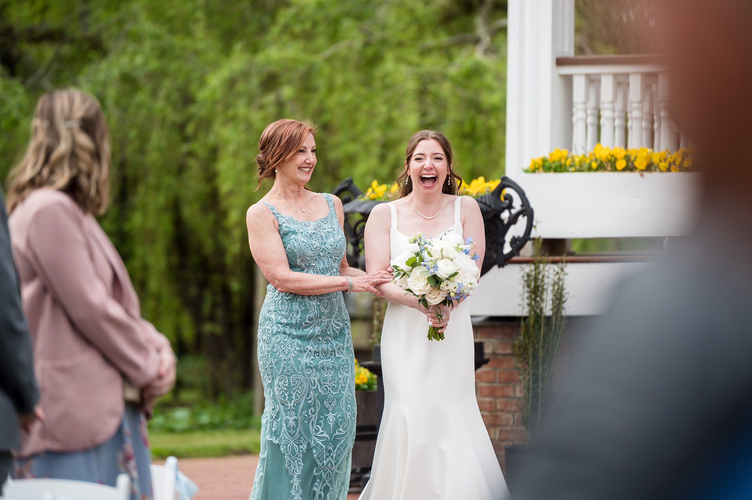 A bride in a white dress and a woman in a teal dress walk down an outdoor aisle, both smiling broadly.