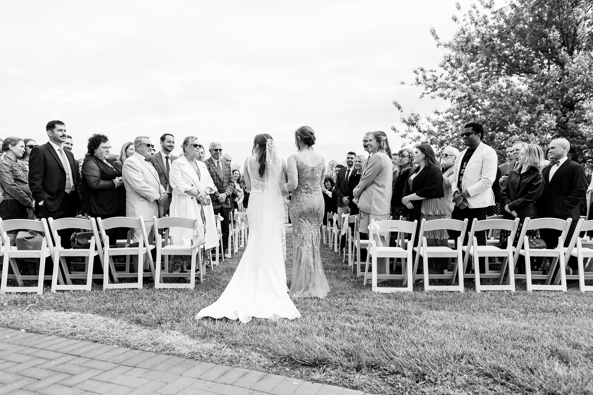 Black and white photo of a bride and another woman walking down an outdoor aisle. Guests are standing and watching, with trees and a cloudy sky in the background.