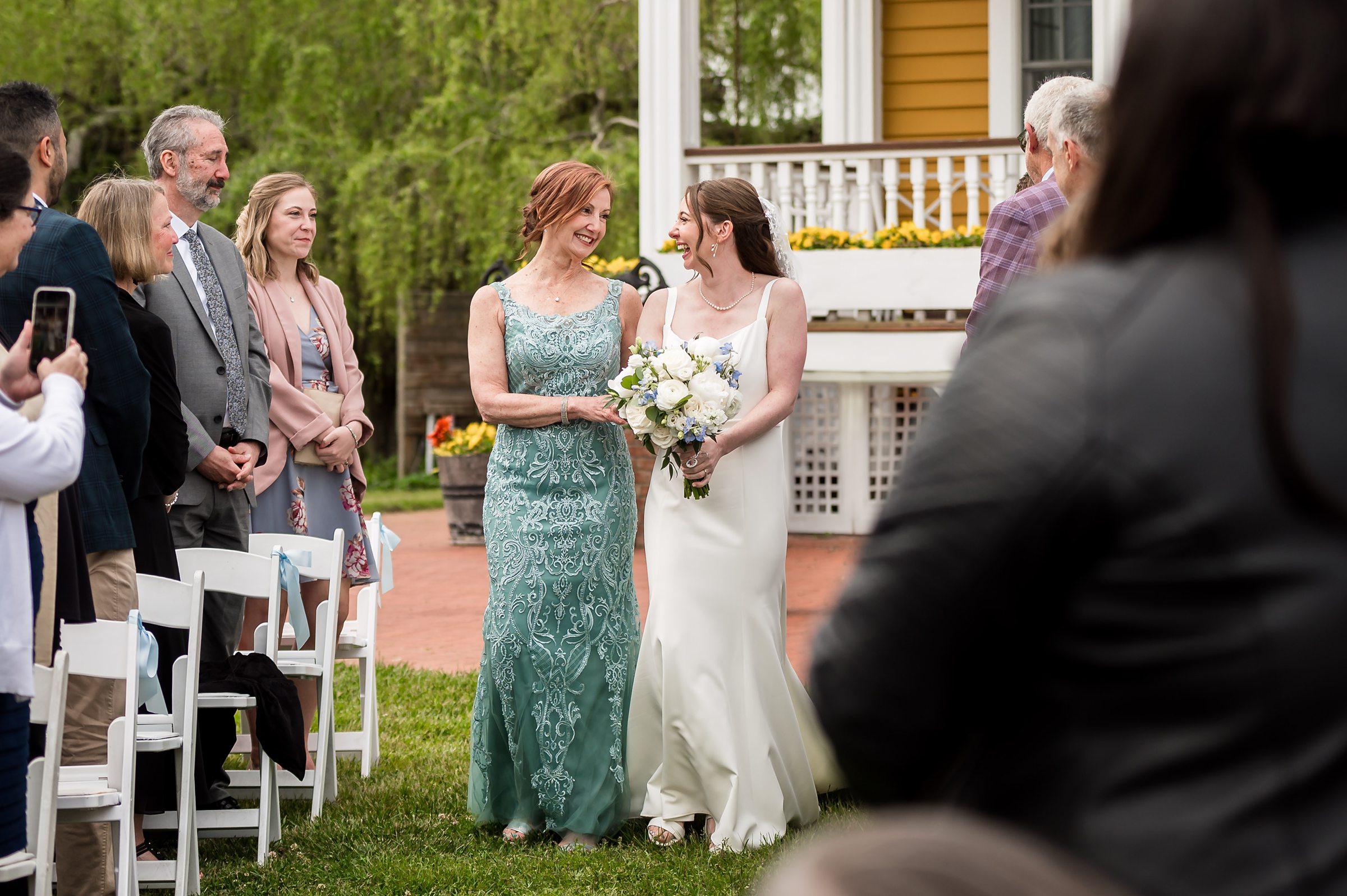 A bride in a white dress holds a bouquet and walks down an outdoor aisle with a woman in a green dress, surrounded by seated and standing guests.