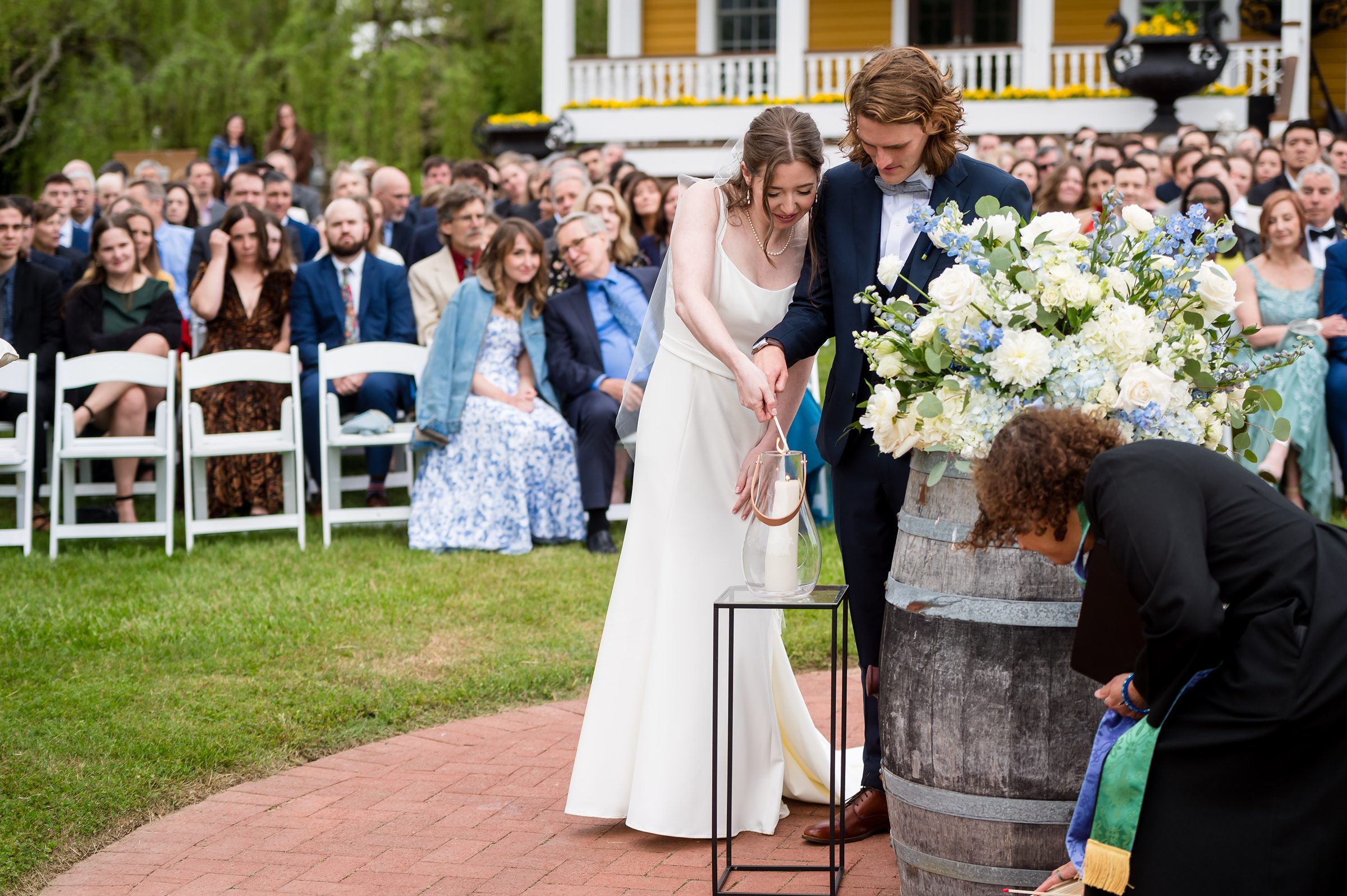 A bride and groom pour liquid into a vase during an outdoor wedding ceremony. Guests are seated in rows in the background, and a person is crouched near a barrel to the right.