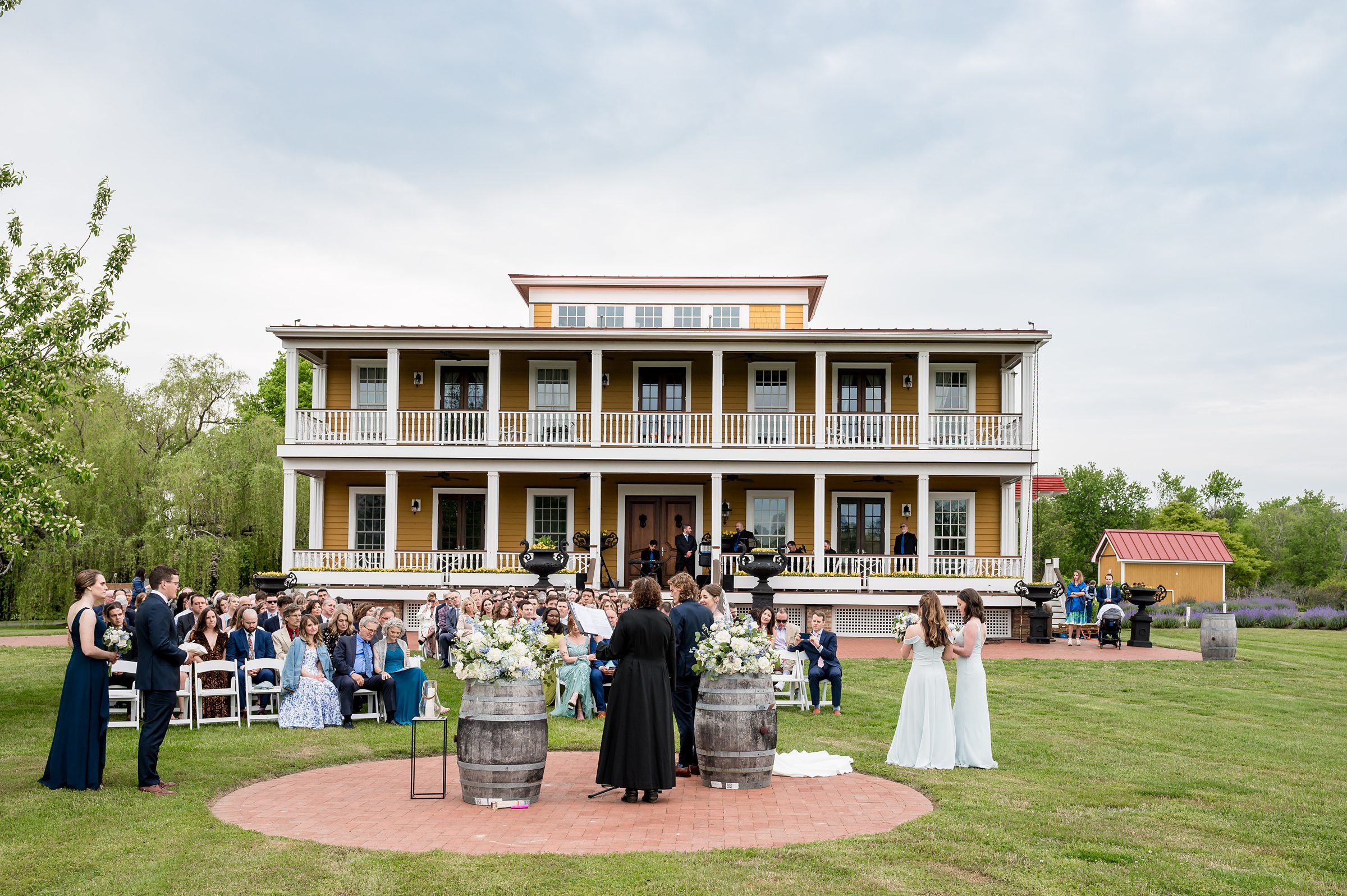 A wedding ceremony takes place outdoors in front of a large, two-story building with a yellow exterior and white railings. Guests are seated on white chairs, and the couple stands with an officiant.