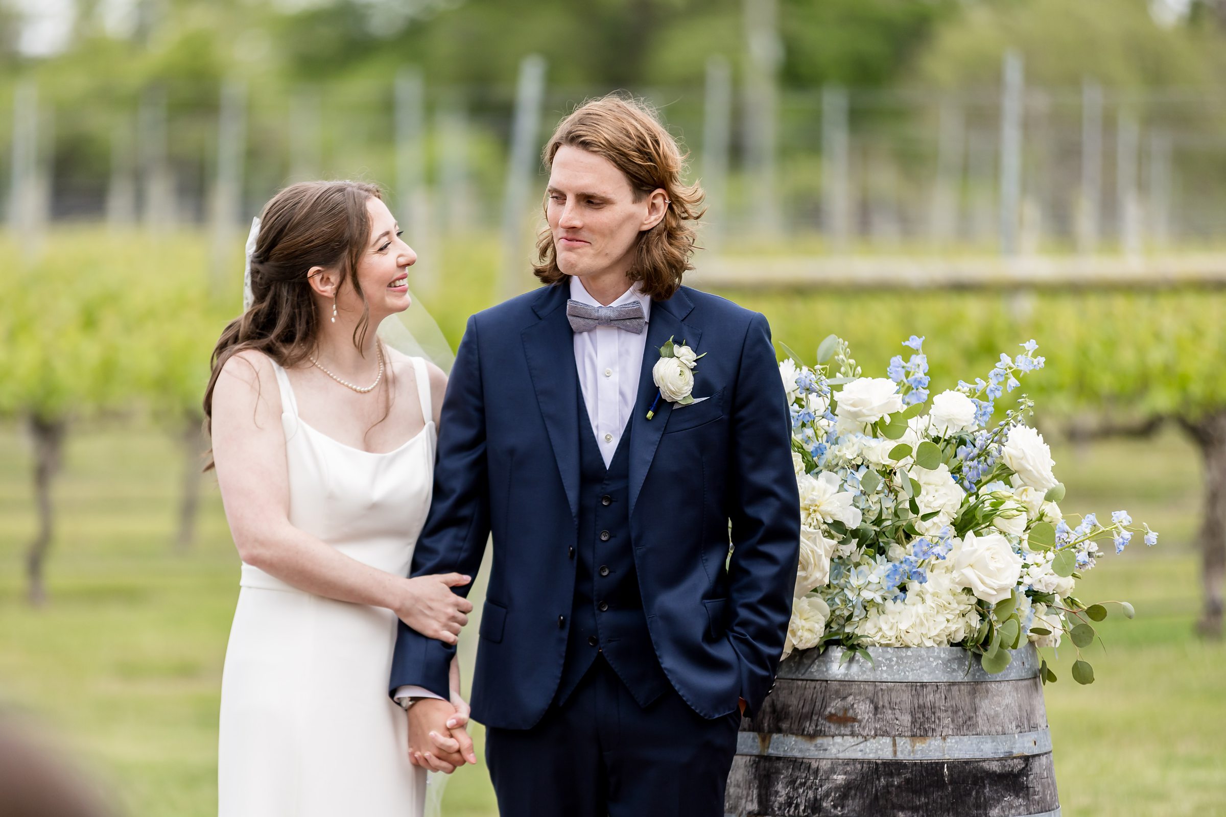 A bride in a white dress and a groom in a navy suit stand together holding hands outdoors, with greenery in the background. The bride smiles at the groom, who looks forward. There are white flowers beside them.