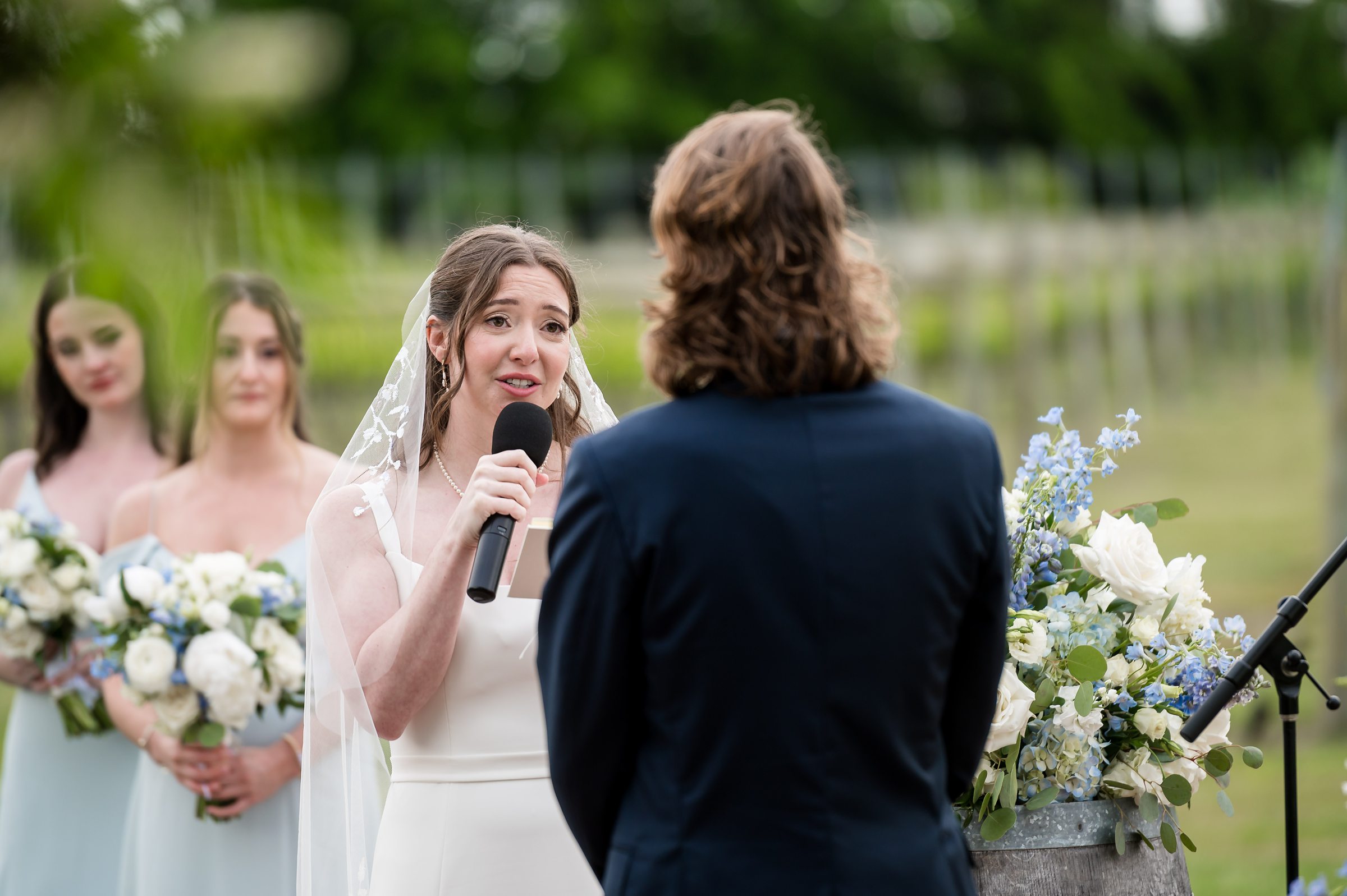 A bride speaks into a microphone during an outdoor wedding ceremony, with bridesmaids holding bouquets standing behind her.