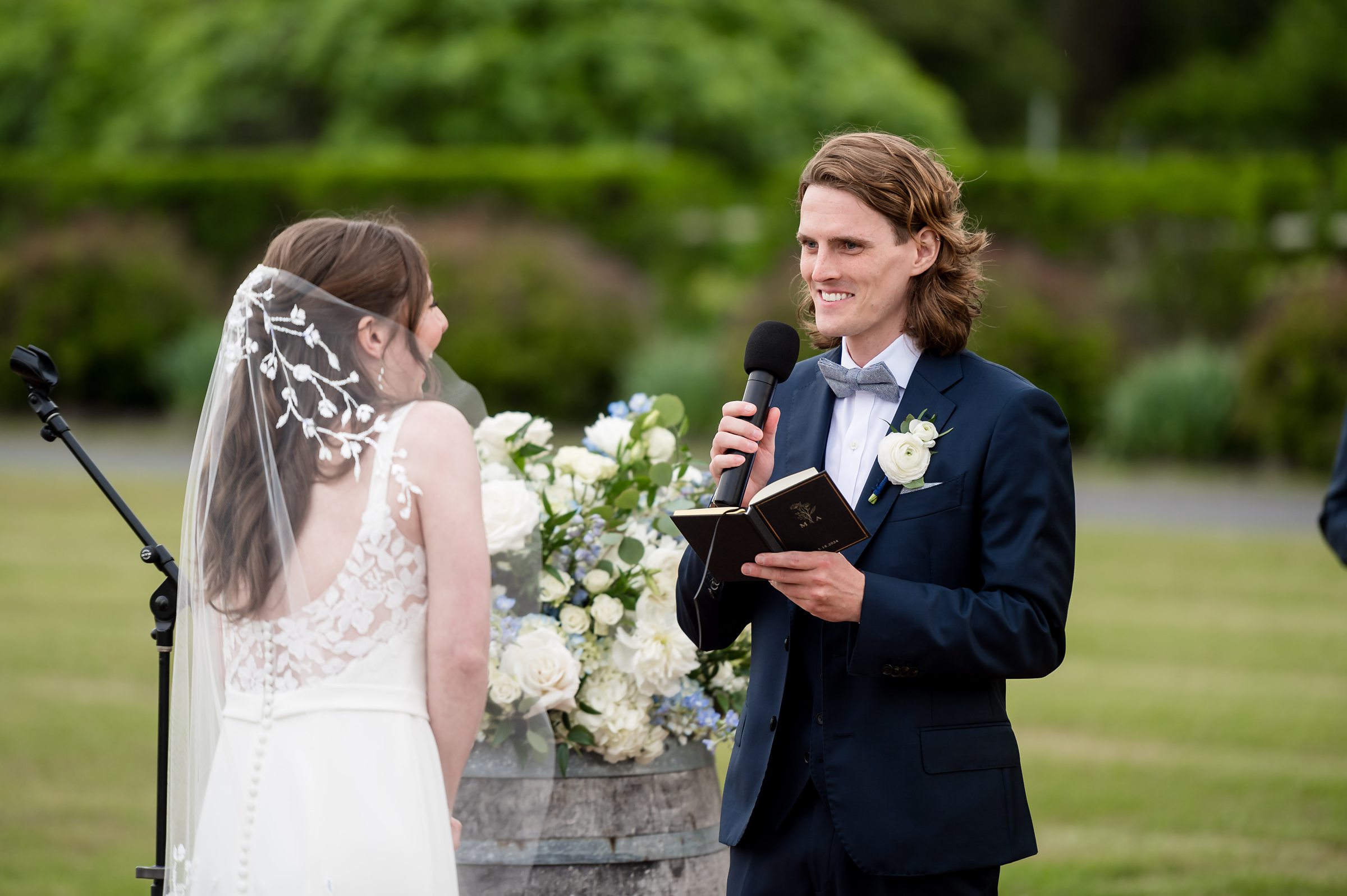 A groom in a navy suit and bow tie reads from a book during an outdoor wedding ceremony. The bride, with a veil and white dress featuring lace details, faces him. Flowers and greenery are in the background.
