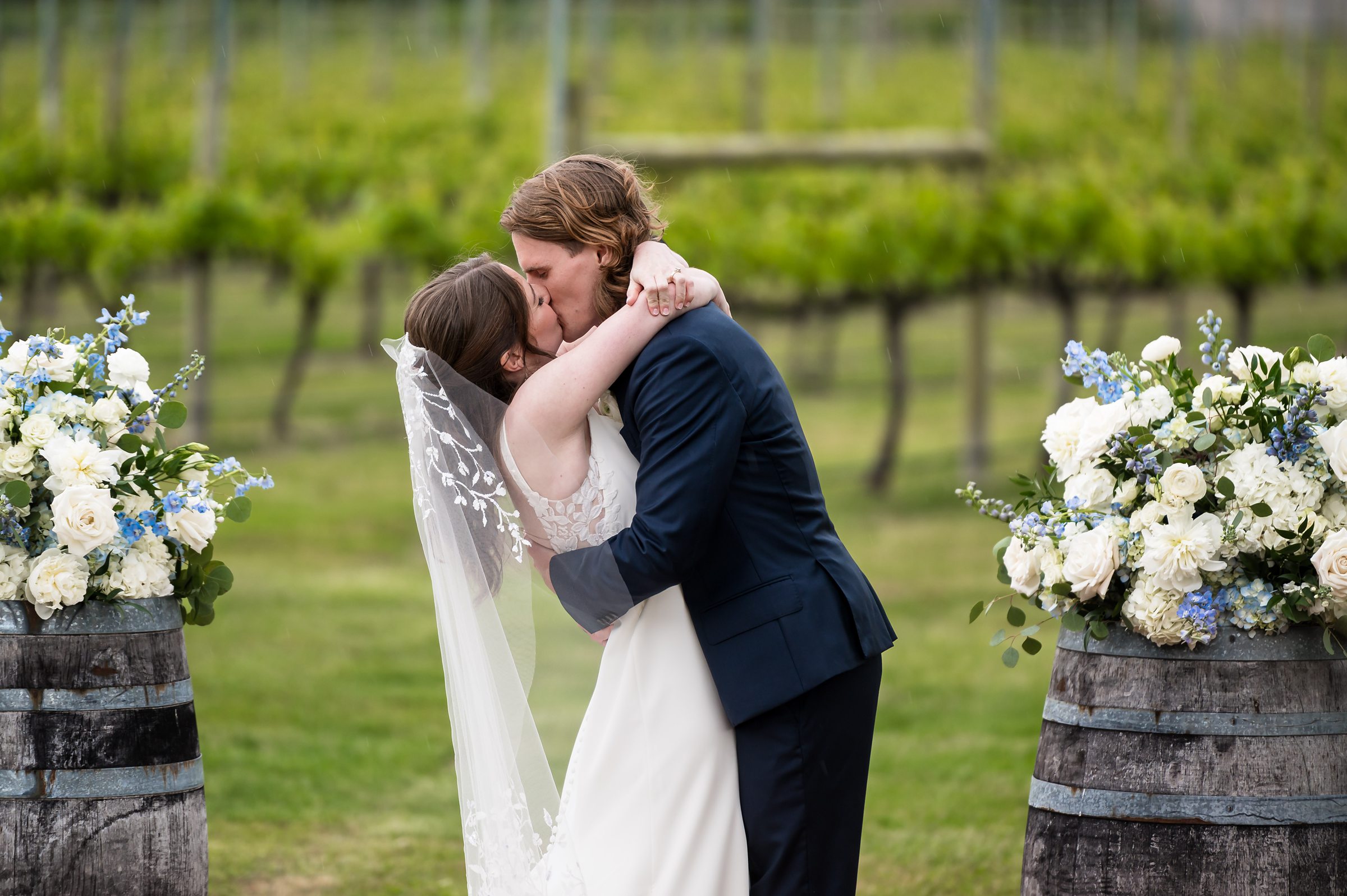 A bride and groom kiss during their outdoor wedding ceremony, surrounded by floral arrangements on wooden barrels and a backdrop of vineyard rows.