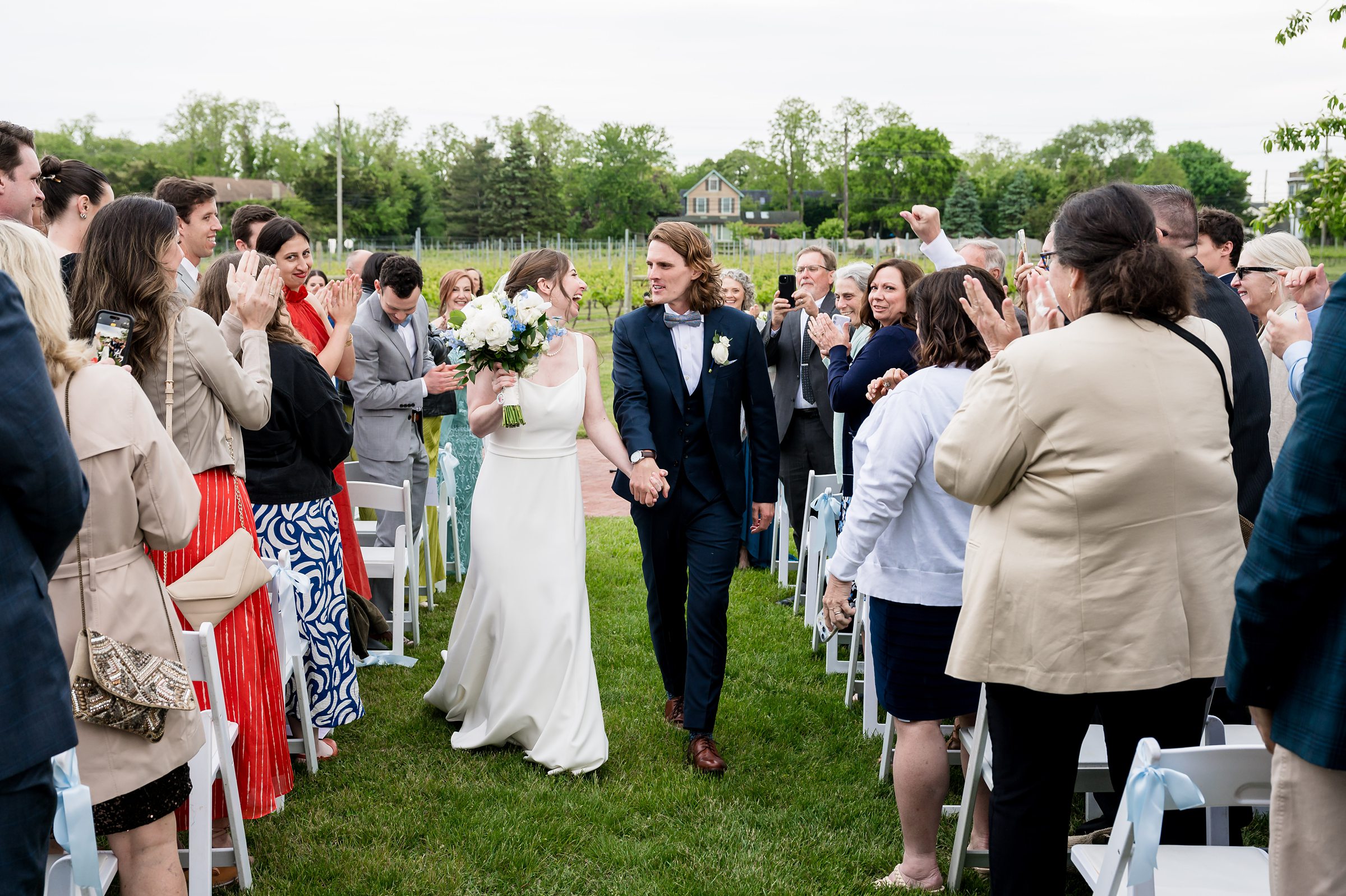 A bride and groom walk down an outdoor aisle holding hands, surrounded by applauding guests. Both are smiling, the groom in a dark suit and the bride in a white gown holding a bouquet.