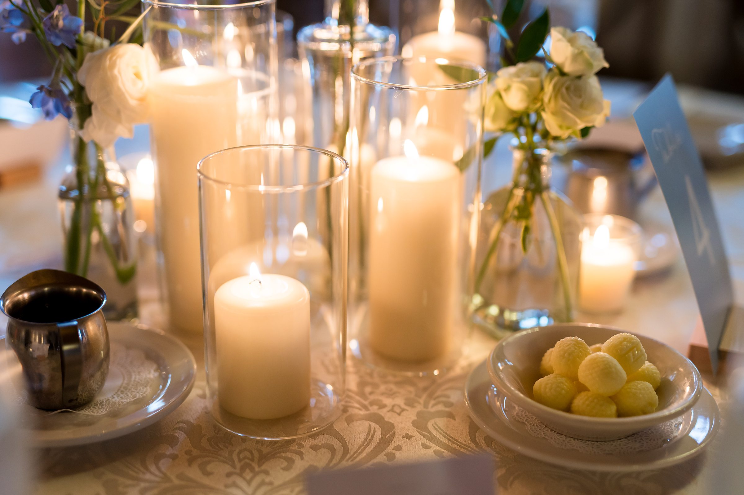 A table centerpiece with lit candles in glass holders, small white flowers in vases, a metal pitcher on a saucer, and a bowl of round yellow candies.