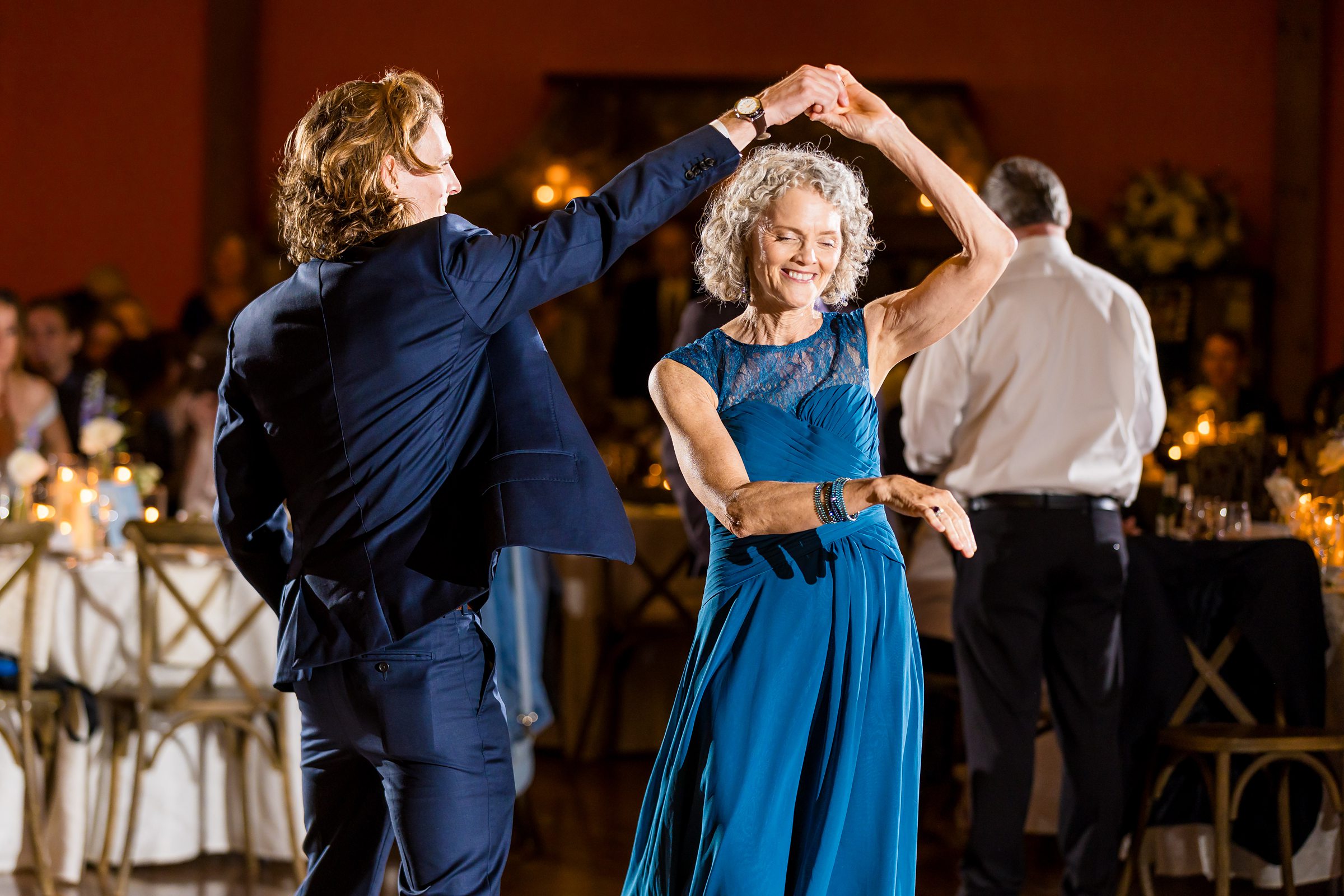 An elderly woman in a blue dress dances and smiles with a man in a blue suit at a formal event with tables and dim lighting in the background.