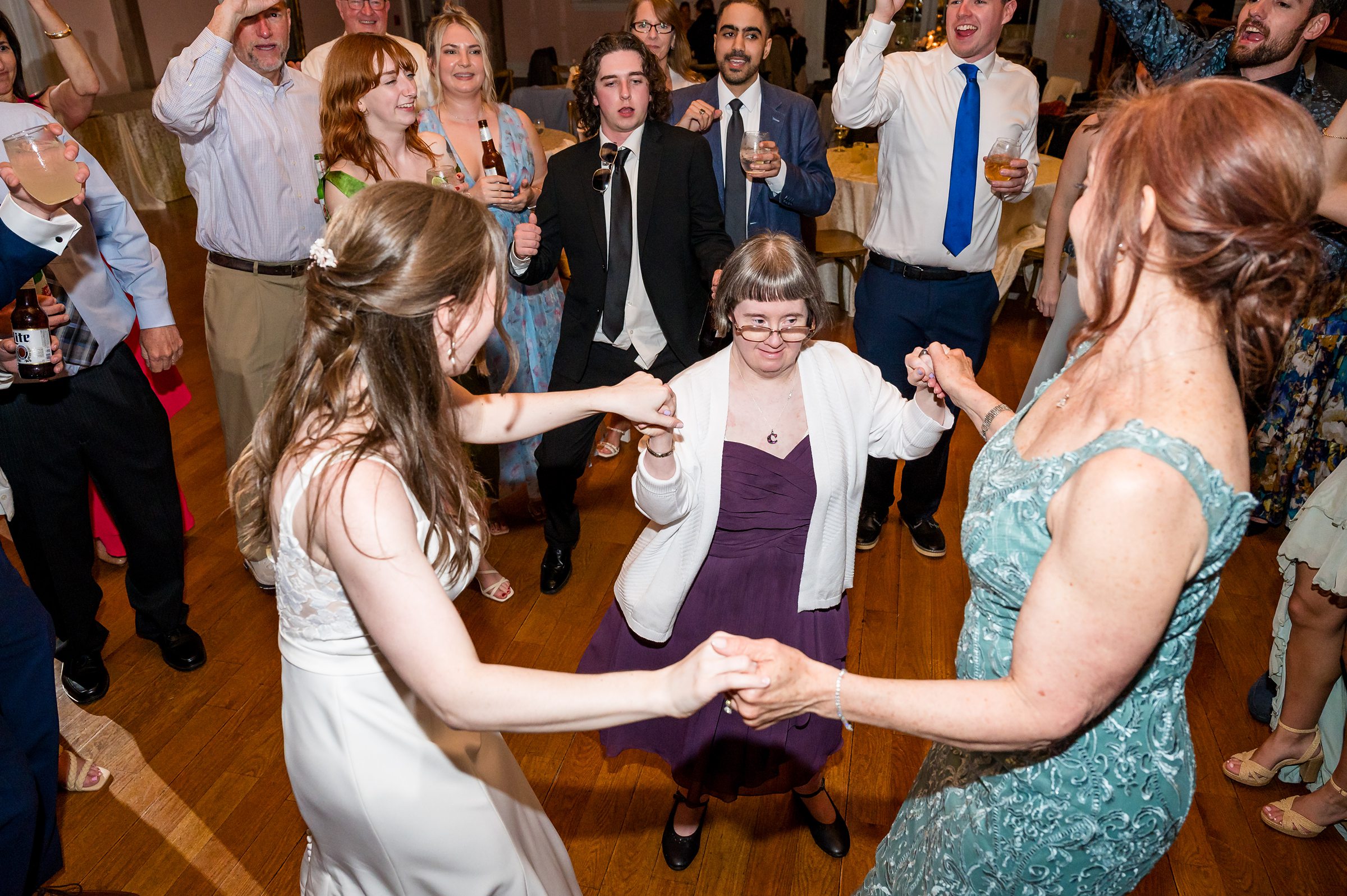 A group of people dressed in formal attire are dancing in a circle at a social event. Some are raising their arms, and three women in the center hold hands, one of them in a white dress.