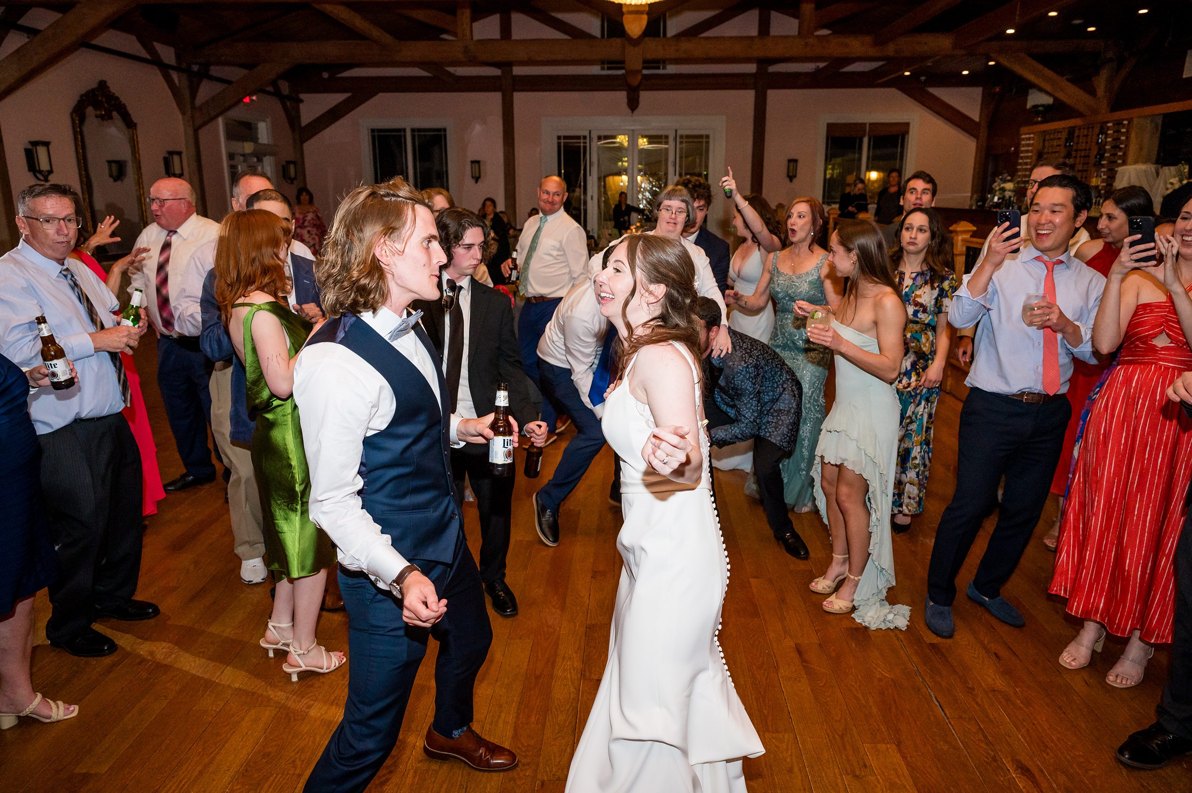 A bride and groom dance together in the center of a lively wedding reception, surrounded by cheering guests on a wooden floor.