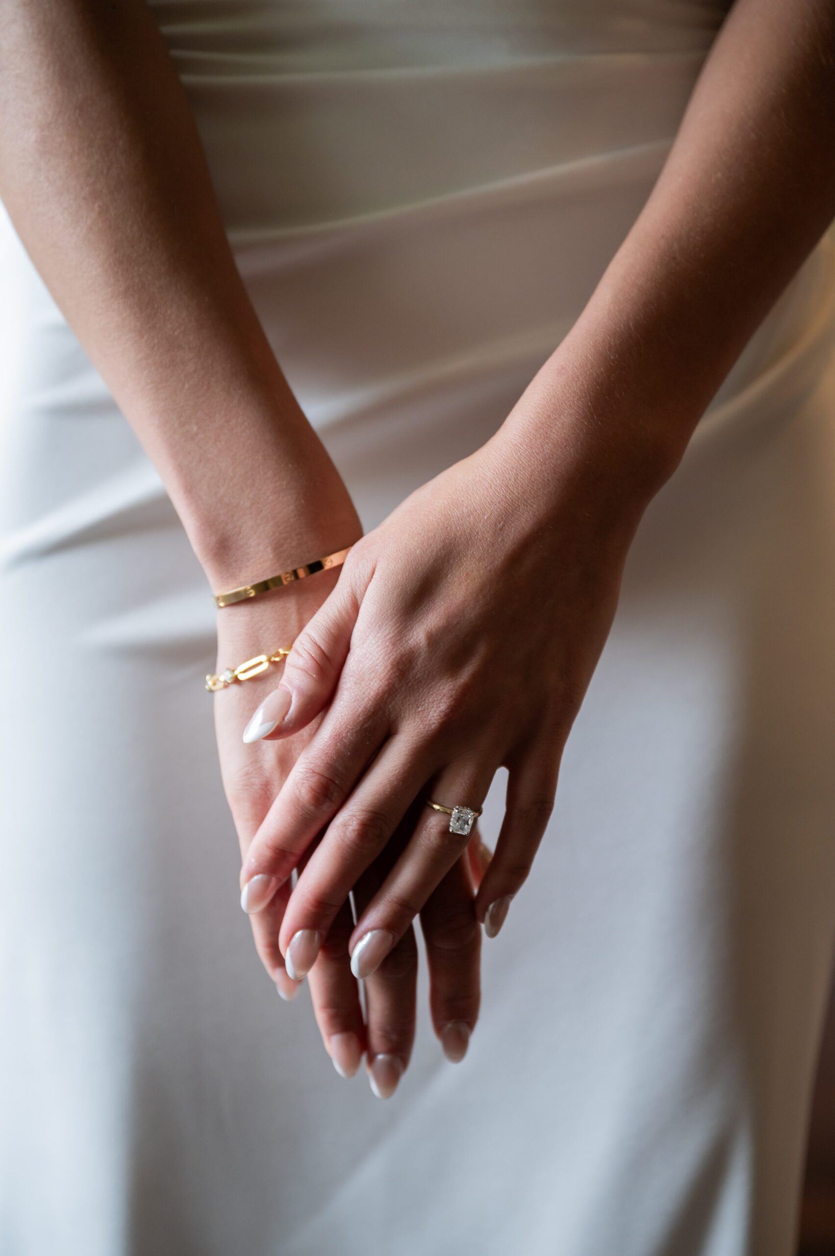 Close-up of a person’s hands with crossed fingers, wearing a gold bracelet and a ring with a square gemstone, set against a white dress.